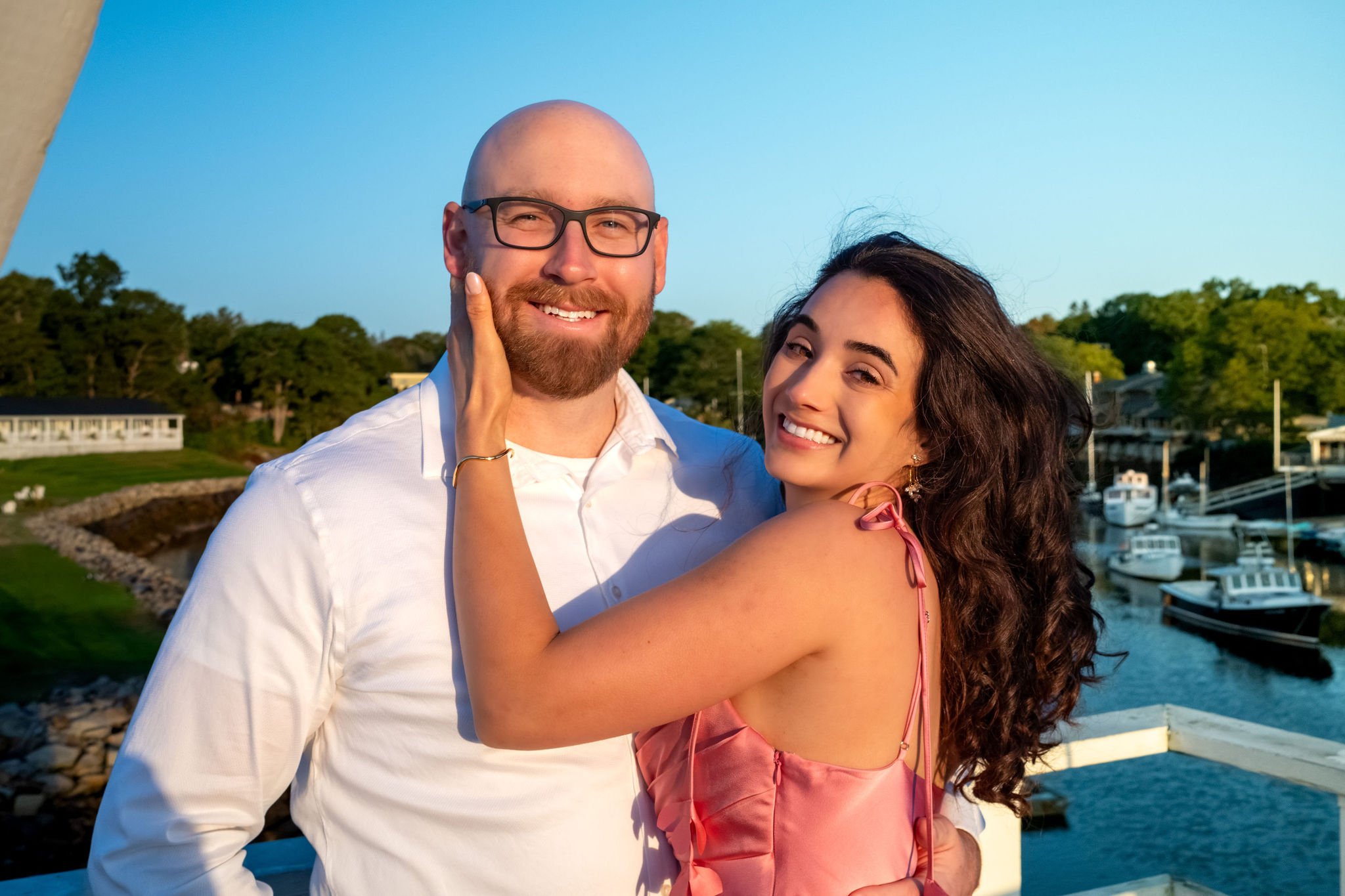 Helena smiling at the camera with her hand on Zack's cheek against a bright blue sky on Marginal Way in Ogunquit Maine sunrise proposal and engagement photography VMG Productions