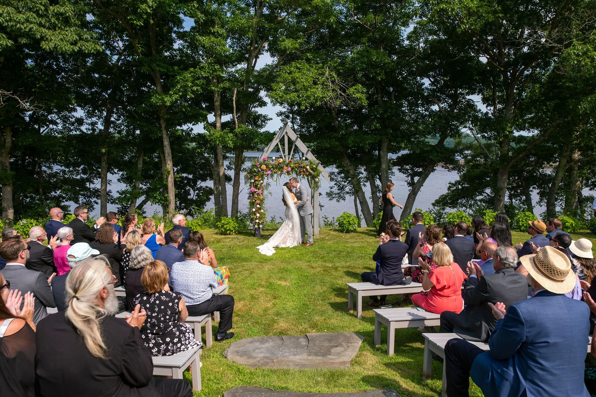 Wide outdoor waterfront ceremony first kiss at Marianmade Farm Wiscasset Maine summer 2024 with guests applauding on wooden benches under tall pine trees with river visible behind colorful floral arch and couple kissing beneath wooden triangle ceremony arbor