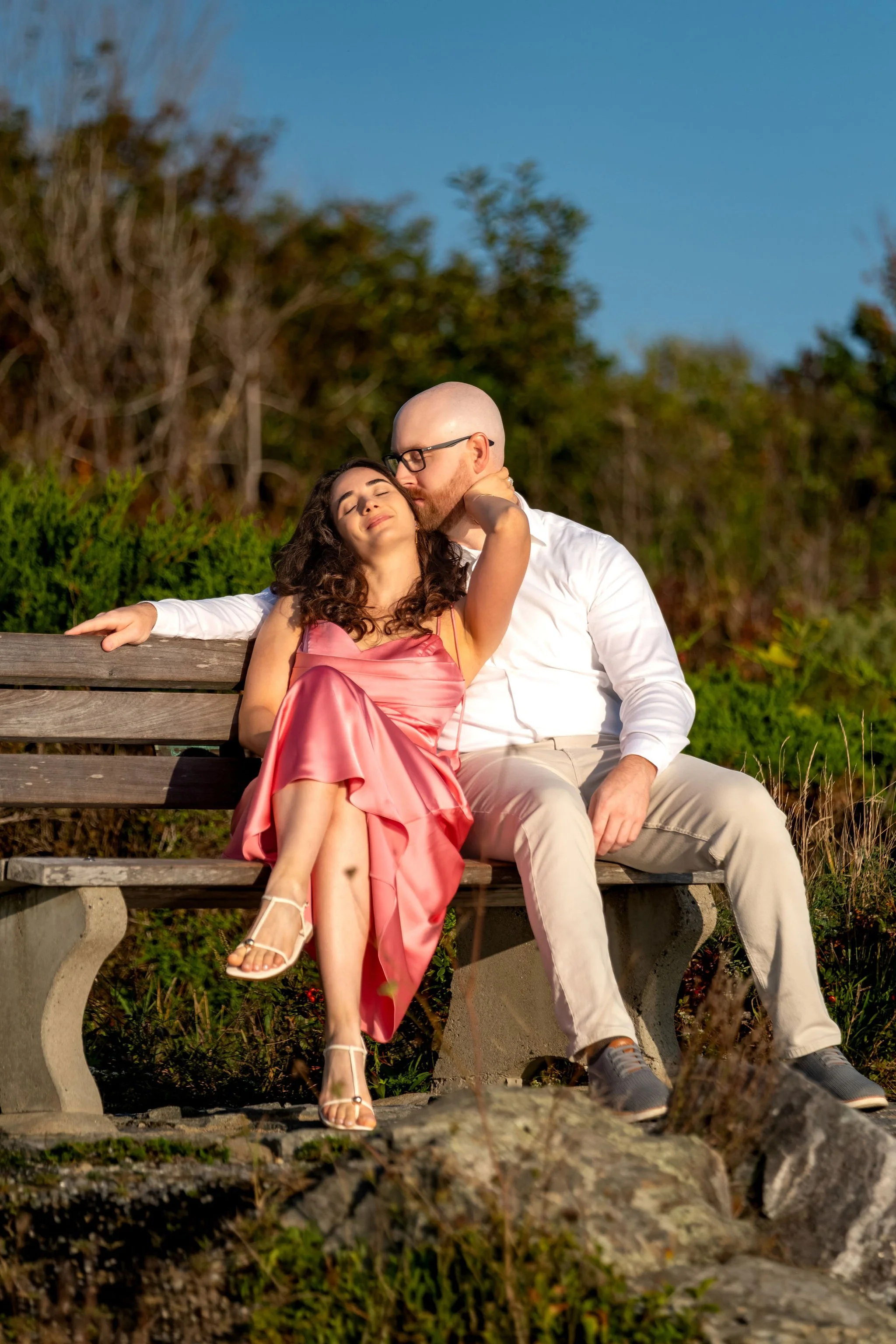 Zack kissing the top of Helena's head as they sit together on the bench on Marginal Way in Ogunquit Maine sunrise proposal and engagement photography VMG Productions