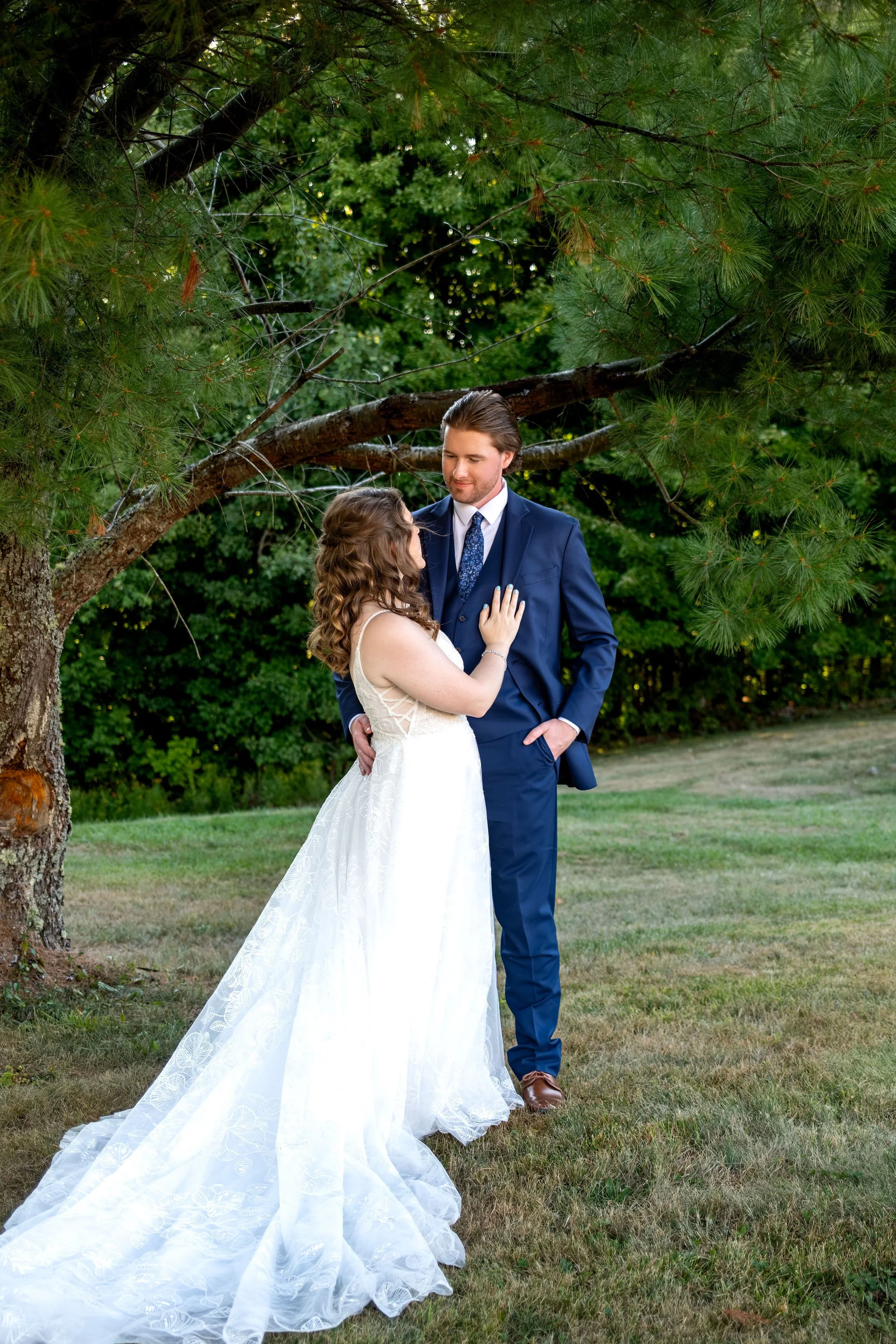 Zachary and Hollie standing close under the branches of a large pine tree with her train on the grass at The Lucerne Inn in Dedham Maine summer wedding photography VMG Productions