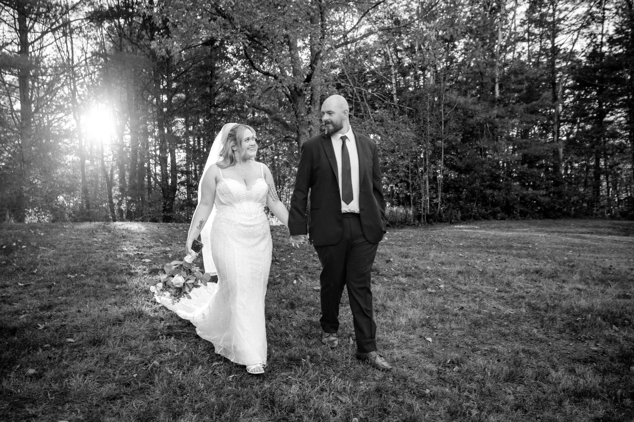Black and white of Ellie and Sam walking hand in hand with the sun flaring through the trees at Boothbay Playhouse in Boothbay Maine fall wedding photography VMG Productions