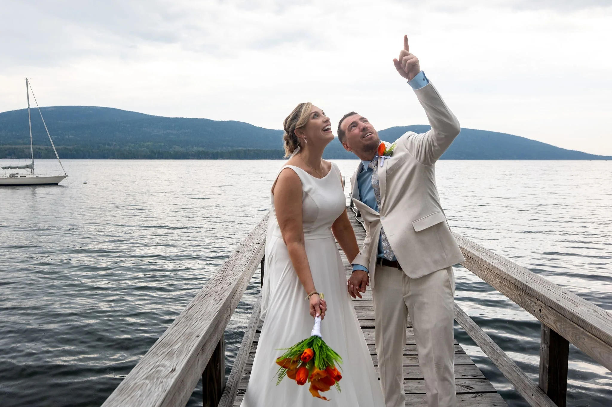 Alli and Adam Baudoin laughing and pointing at the sky from the dock on Somes Sound with a sailboat and the Acadia hills behind them in Mount Desert Maine spring wedding photography VMG Productions
