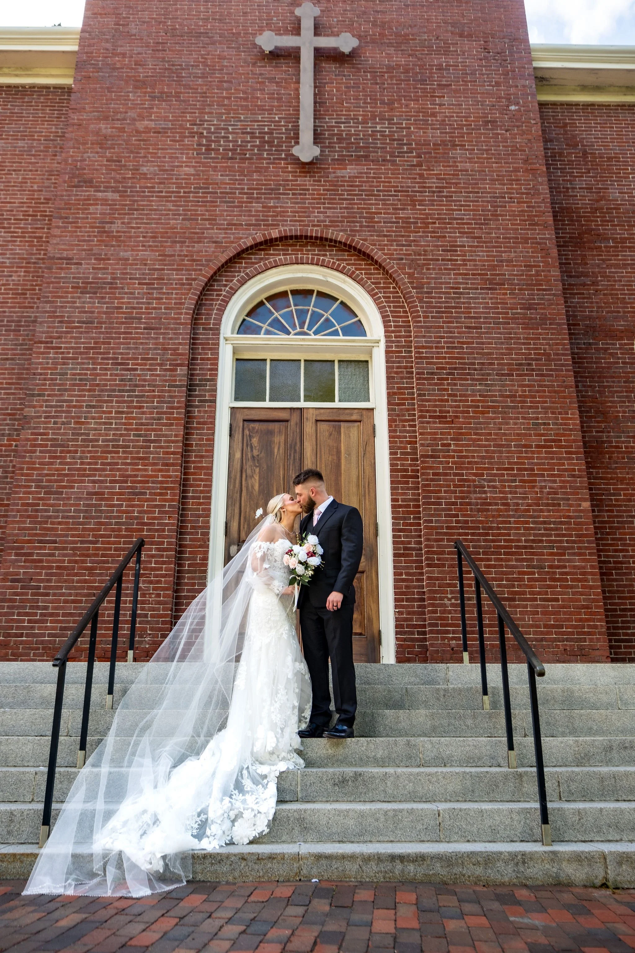 Jeremiah and Vienna wide vertical composition showing church facade and cross above couple at Holy Trinity Greek Orthodox Church Portland Maine spring wedding photography VMG Productions