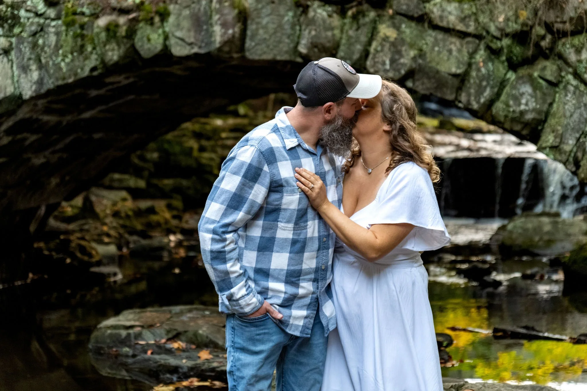 Alison and Dave kissing beneath the granite arch with the brook tumbling over the rocks behind them at Vaughan Woods in Hallowell Maine fall engagement photography VMG Productions