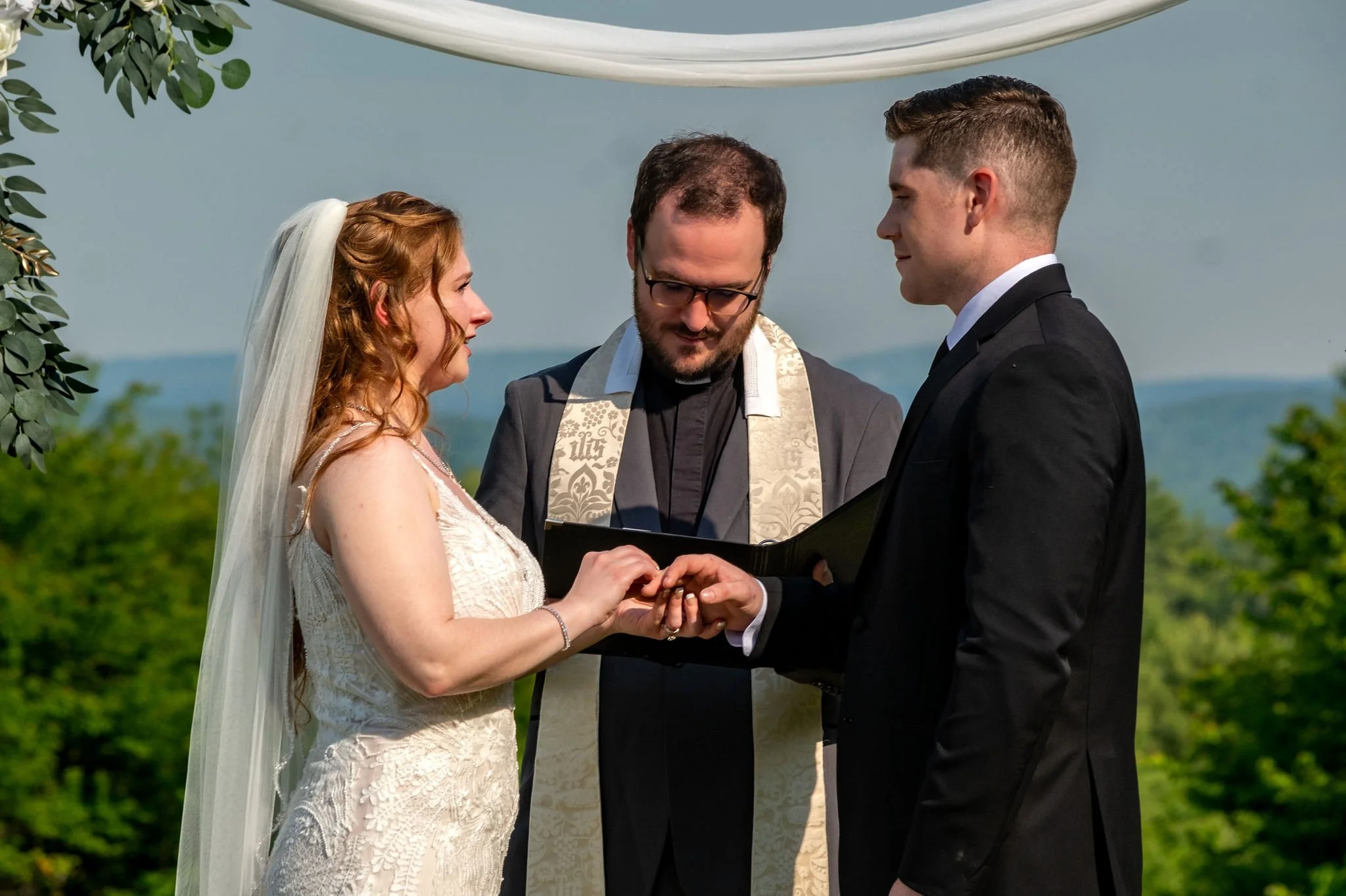 Kayla and Ethan DeAngelis ring exchange under the floral arch at their Sanbornton New Hampshire backyard wedding photography VMG Productions