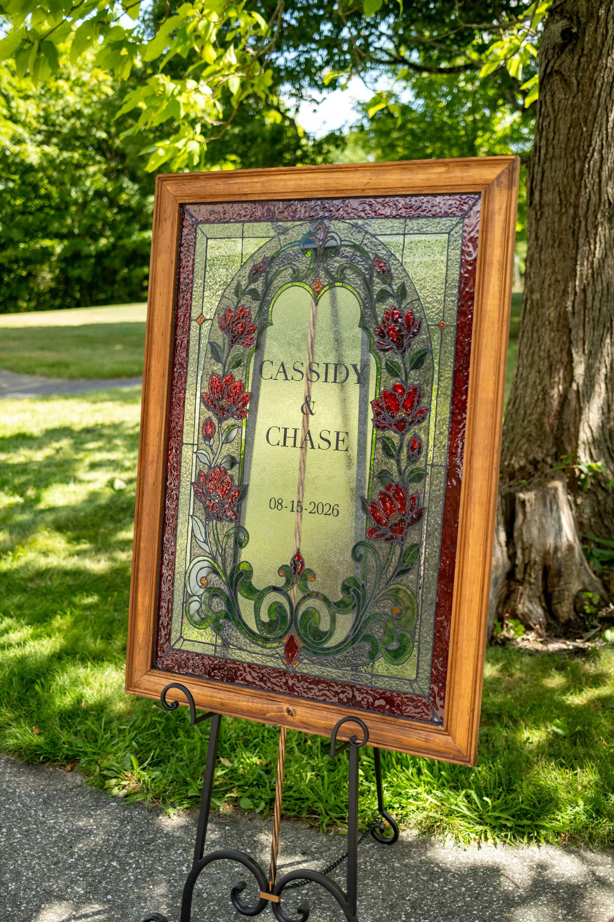 Stained glass welcome sign with Cassidy and Chase's names on an easel under the maples at Maple Hill Farm Inn and Conference Center in Hallowell Maine summer wedding photography VMG Productions