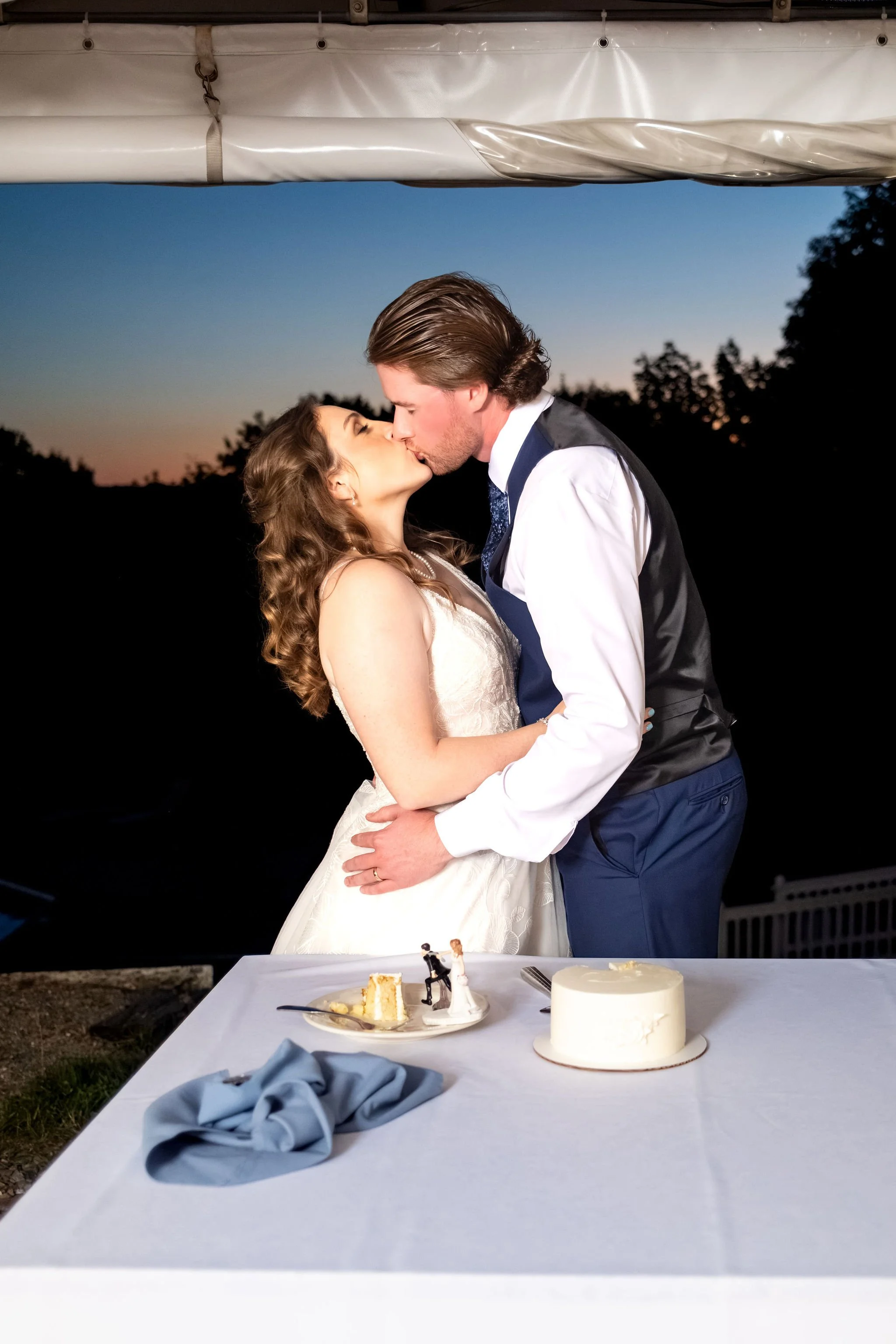 Zachary and Hollie kissing over the cake table at dusk at The Lucerne Inn in Dedham Maine summer wedding photography VMG Productions