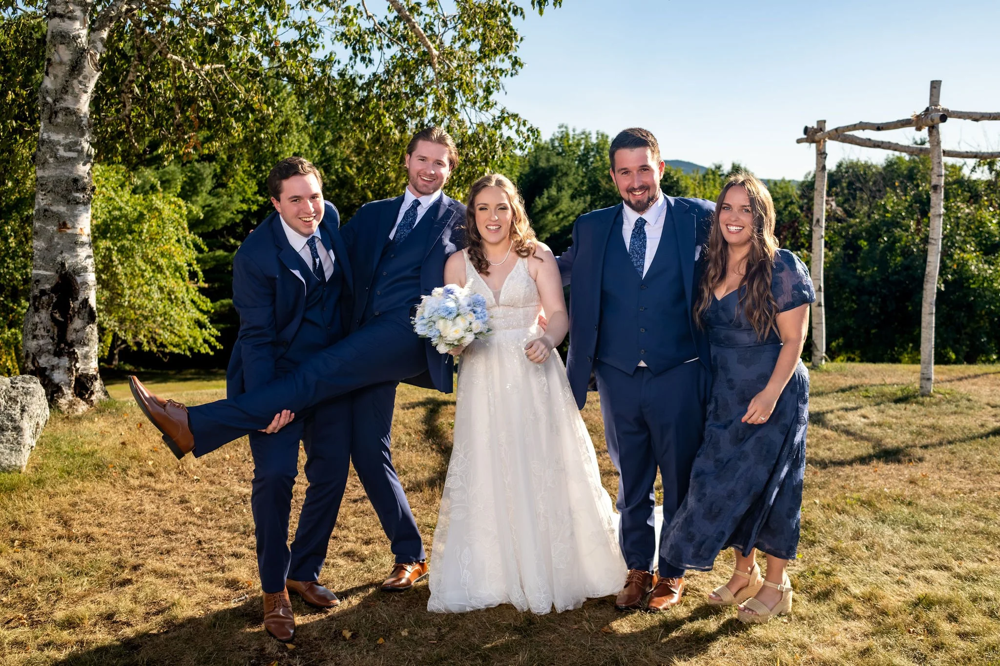 Zachary and Hollie laughing with members of their wedding party as one groomsman kicks his leg in the air at The Lucerne Inn in Dedham Maine summer wedding photography VMG Productions