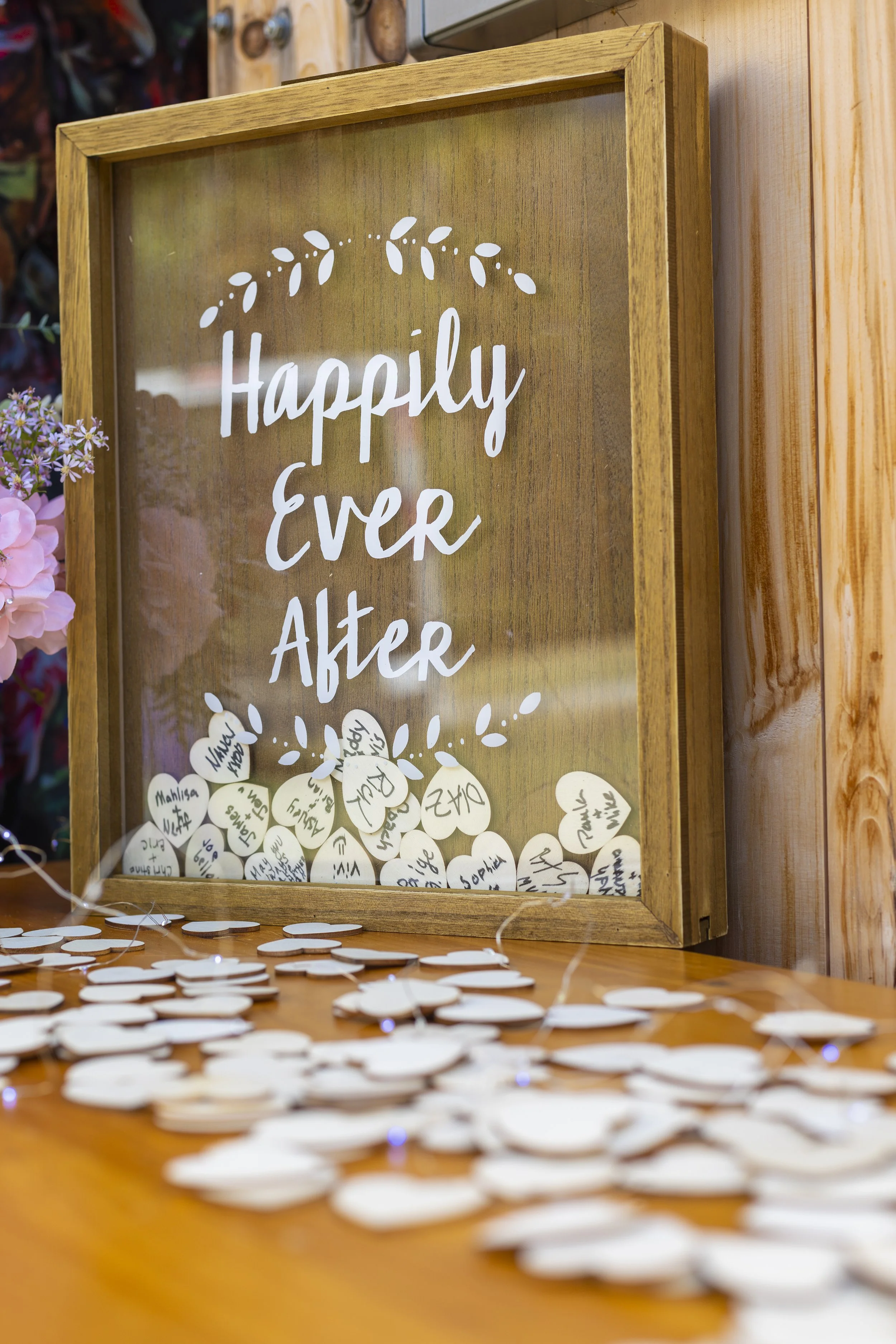 Close up of wooden heart guest tokens filling Happily Ever After shadow box frame with handwritten guest names and wishes at Harvest Moon Farm Winthrop Maine barn wedding reception