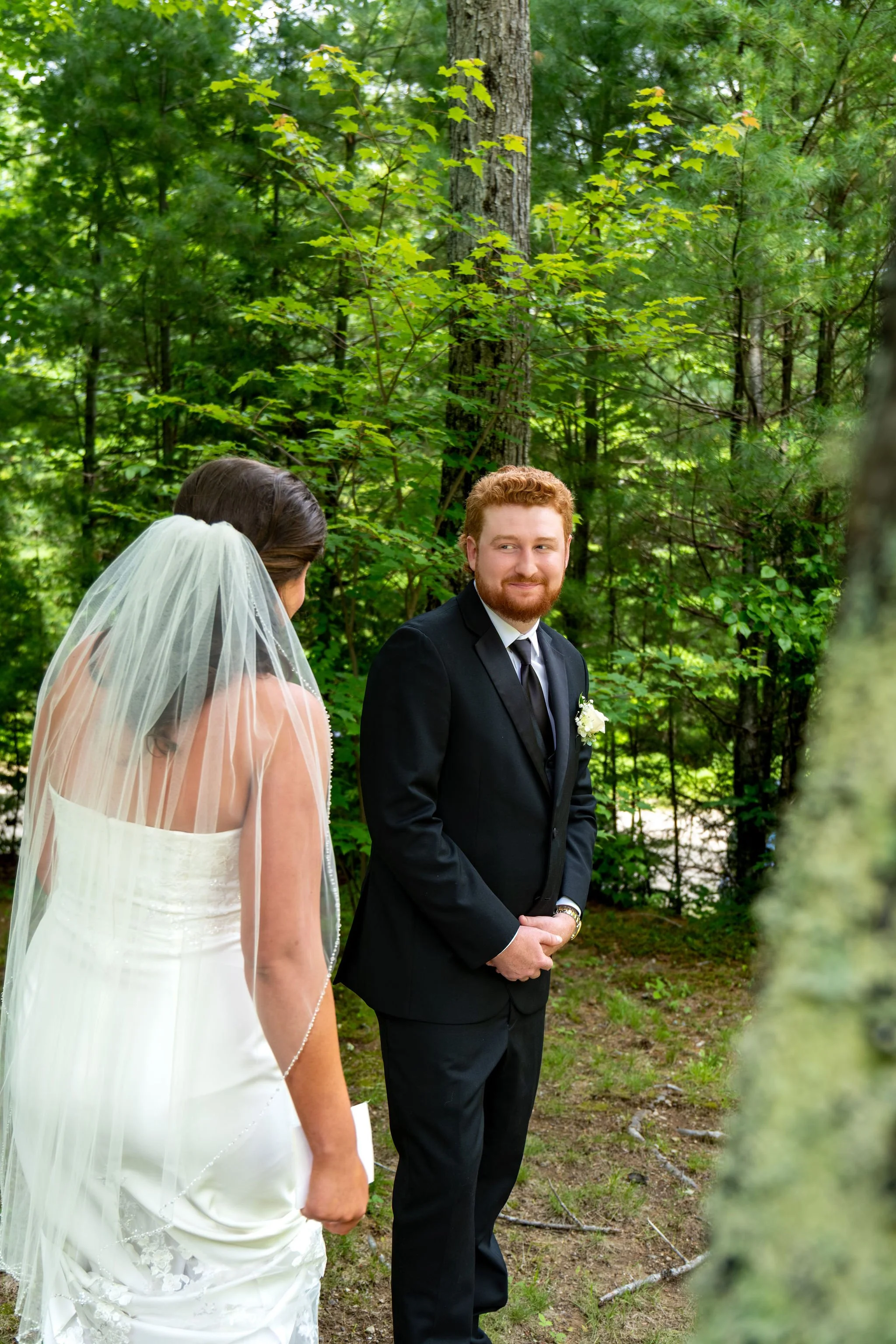Alyssa and Colby the groom smiling at his bride during their first look among the pines in Bethel Maine summer wedding photography VMG Productions