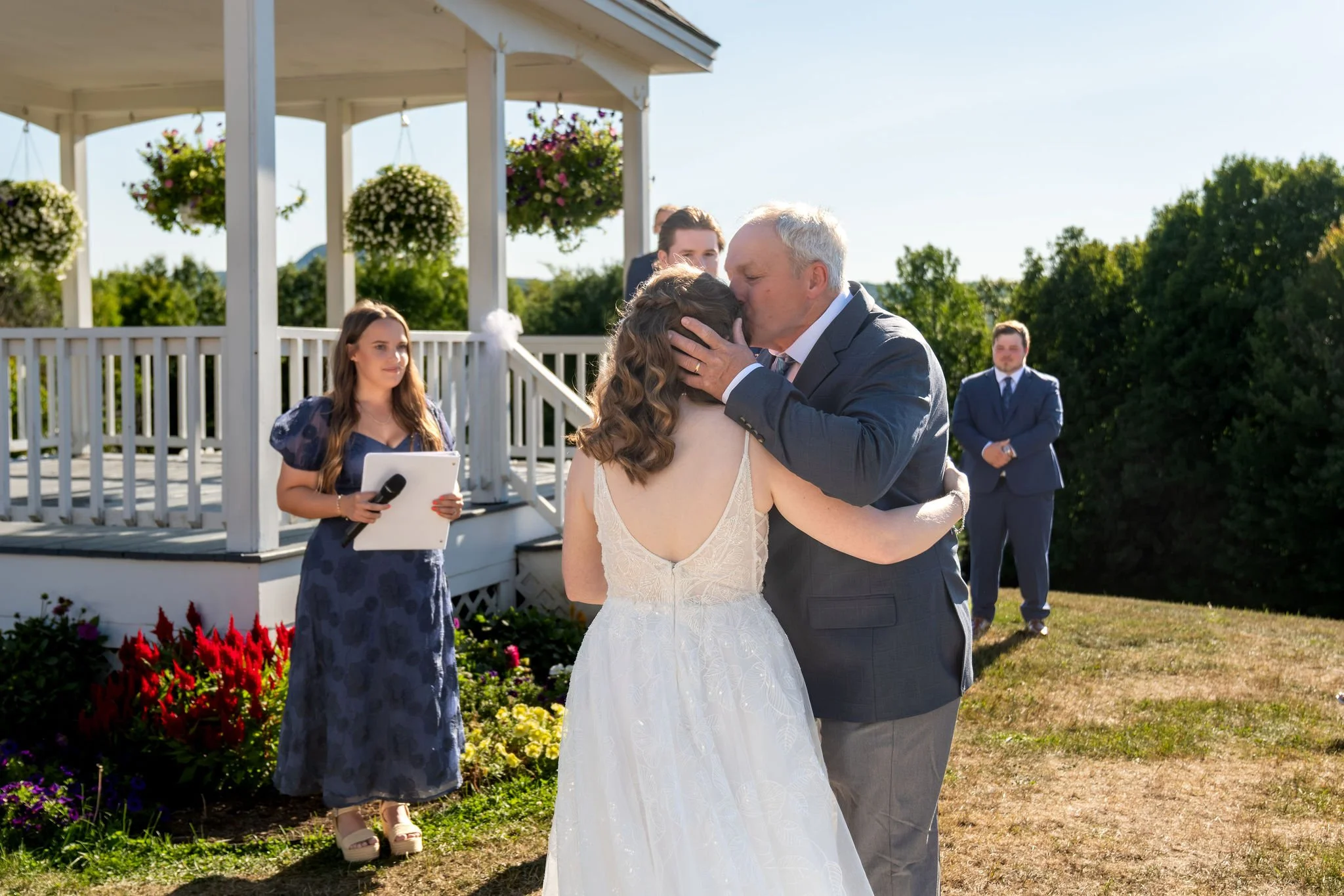 Hollie's dad kissing her on the cheek as he gives her away at the gazebo steps while Zachary and the officiant look on at The Lucerne Inn in Dedham Maine summer wedding photography VMG Productions