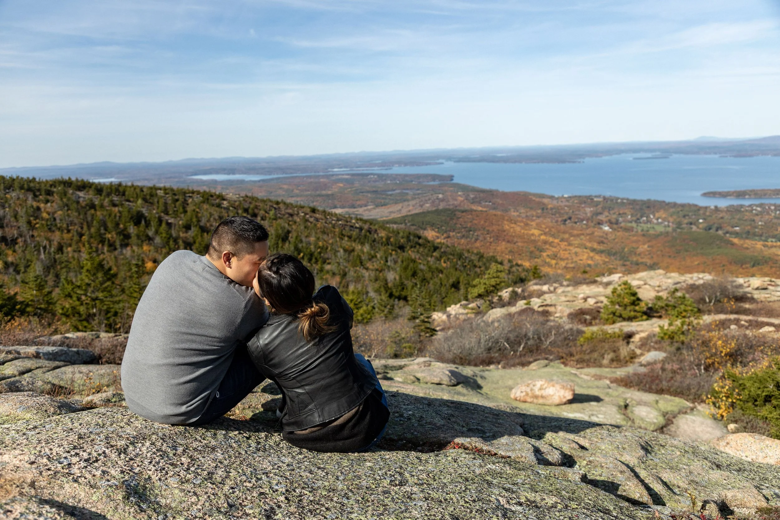 Agnes and Peter from behind sitting on summit rocks looking out at fall foliage and Frenchman Bay Acadia National Park engagement VMG Productions