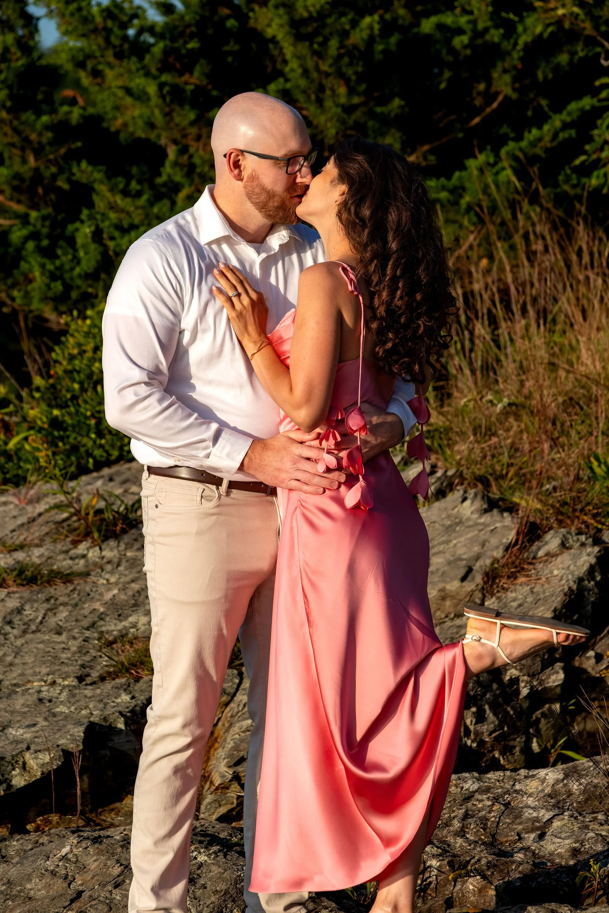Zack and Helena kissing on the rocks with the golden light on the grasses behind them on Marginal Way in Ogunquit Maine sunrise proposal and engagement photography VMG Productions