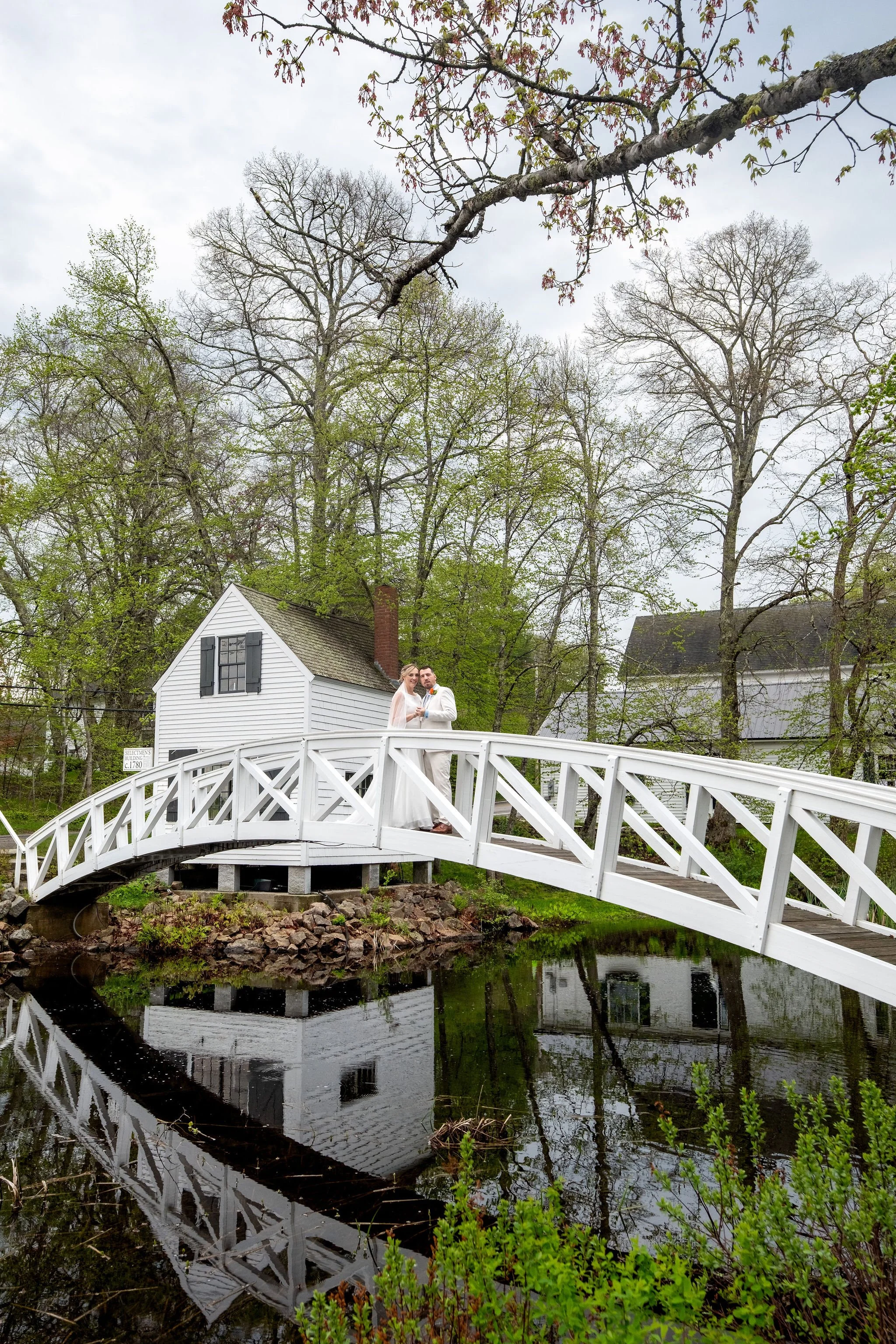 Alli and Adam Baudoin standing on the white arched Somesville footbridge with its reflection in the mill pond and the historic white village buildings behind them on Mount Desert Island Maine spring wedding photography VMG Productions