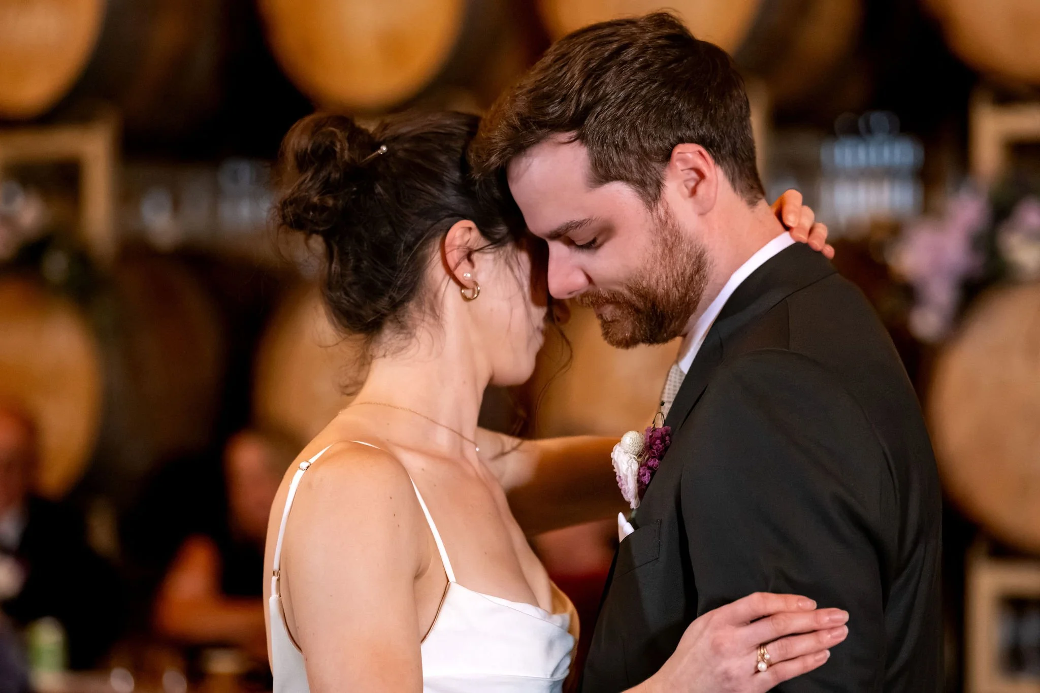 Victoria and Jack dancing with their foreheads together with warm barrels glowing behind them at The Cellars at Allagash Brewing Company in Portland Maine fall wedding photography VMG Productions