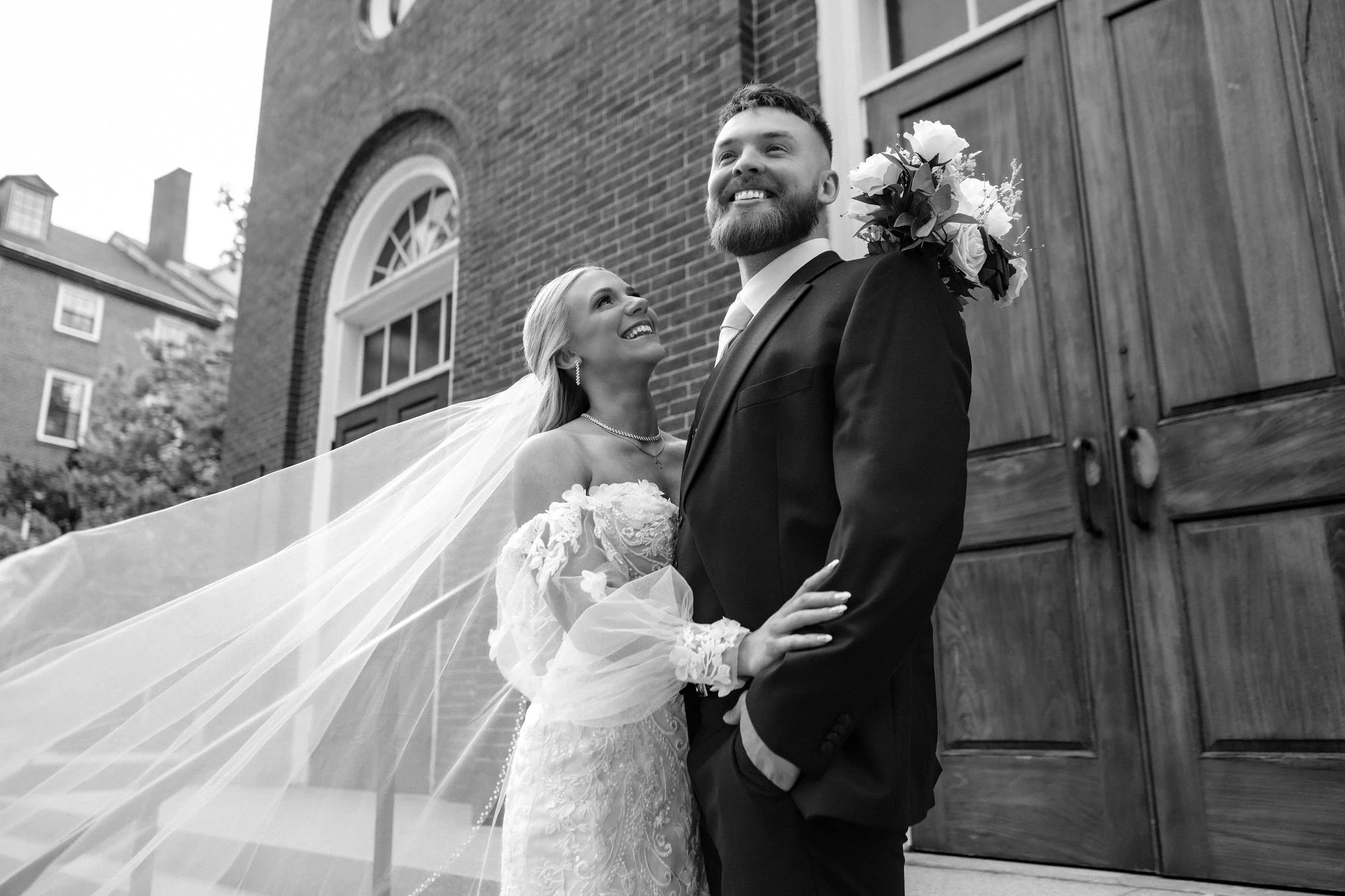 Jeremiah and Vienna black and white portrait of the couple outside the church with the veil sweeping dramatically across the frame at Holy Trinity Greek Orthodox Church Portland Maine spring wedding photography VMG Productions