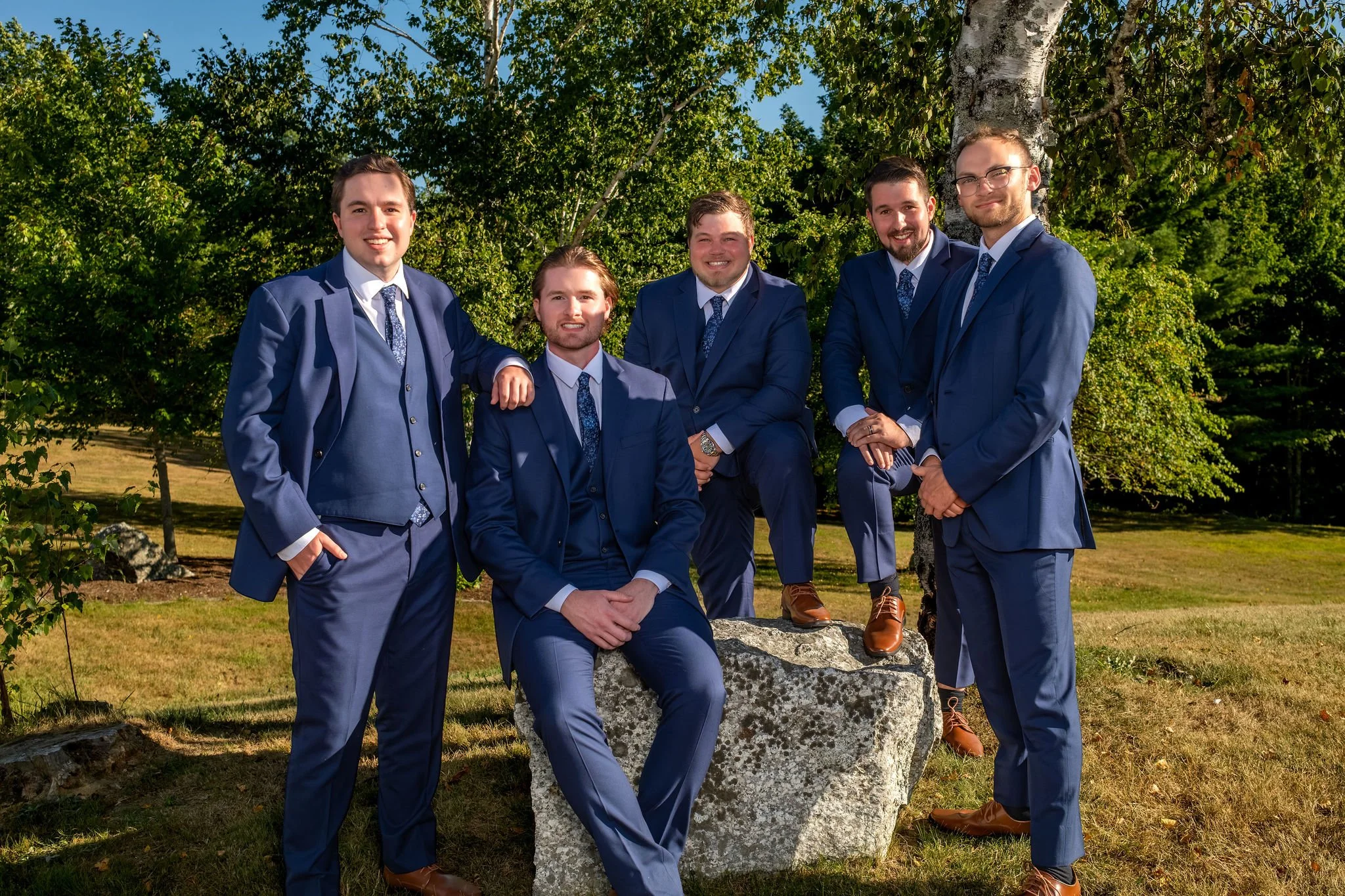 Zachary with his groomsmen in navy suits gathered around a granite boulder at The Lucerne Inn in Dedham Maine summer wedding photography VMG Productions