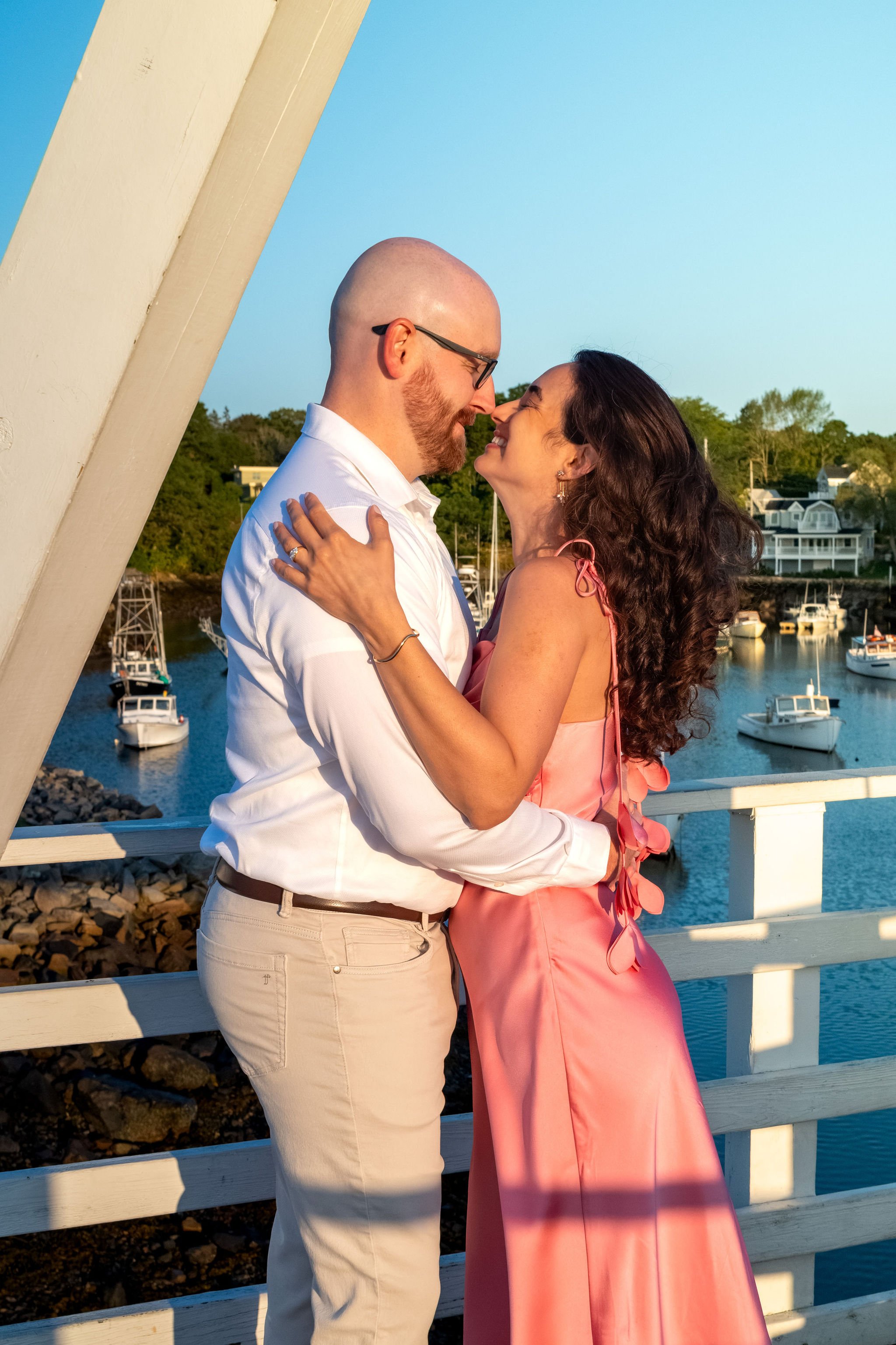 Zack and Helena hugging with their foreheads together on the footbridge as the harbor glows below on Marginal Way in Ogunquit Maine sunrise proposal and engagement photography VMG Productions
