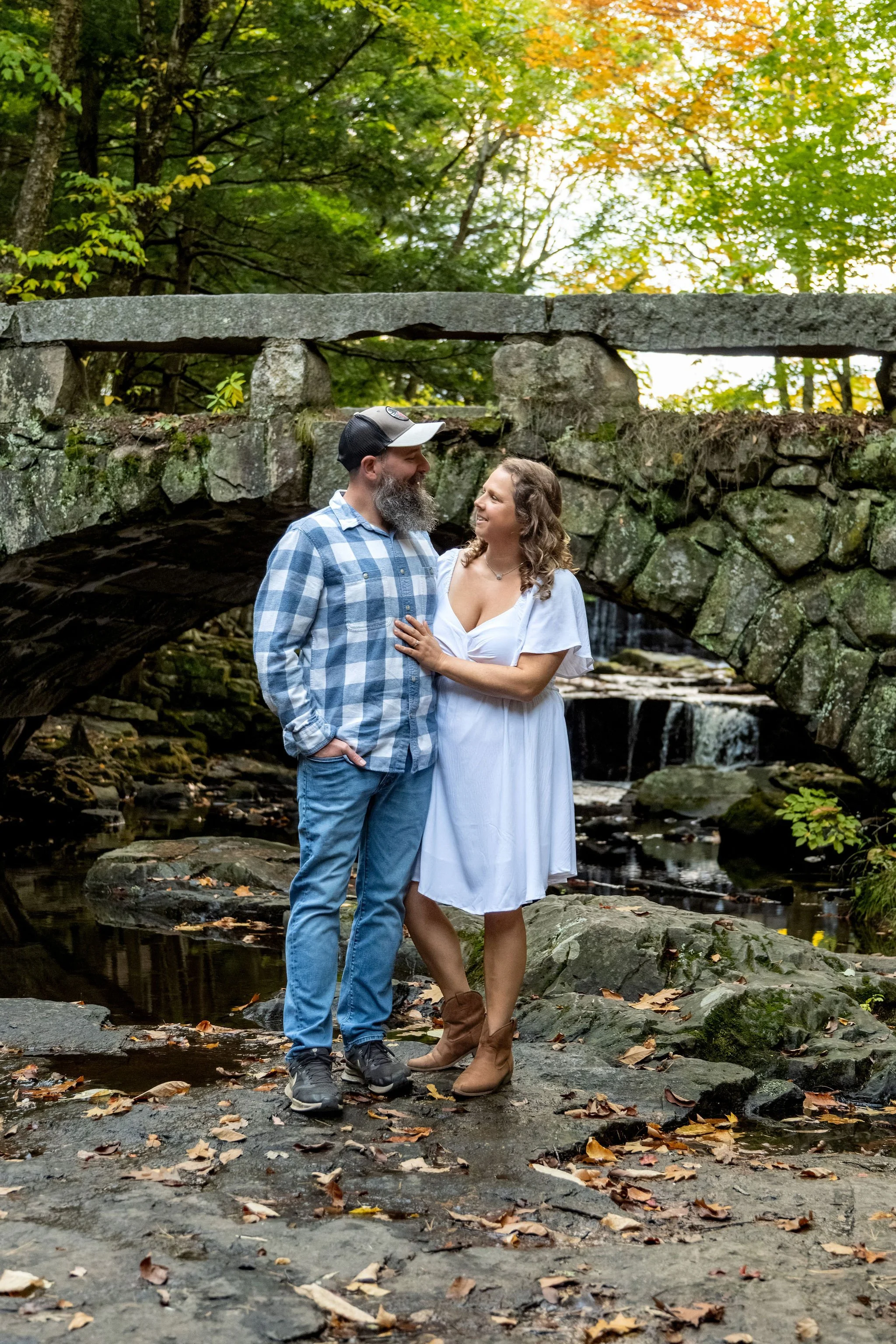 Alison and Dave looking at each other in front of the arch bridge with fall color in the trees above at Vaughan Woods in Hallowell Maine fall engagement photography VMG Productions