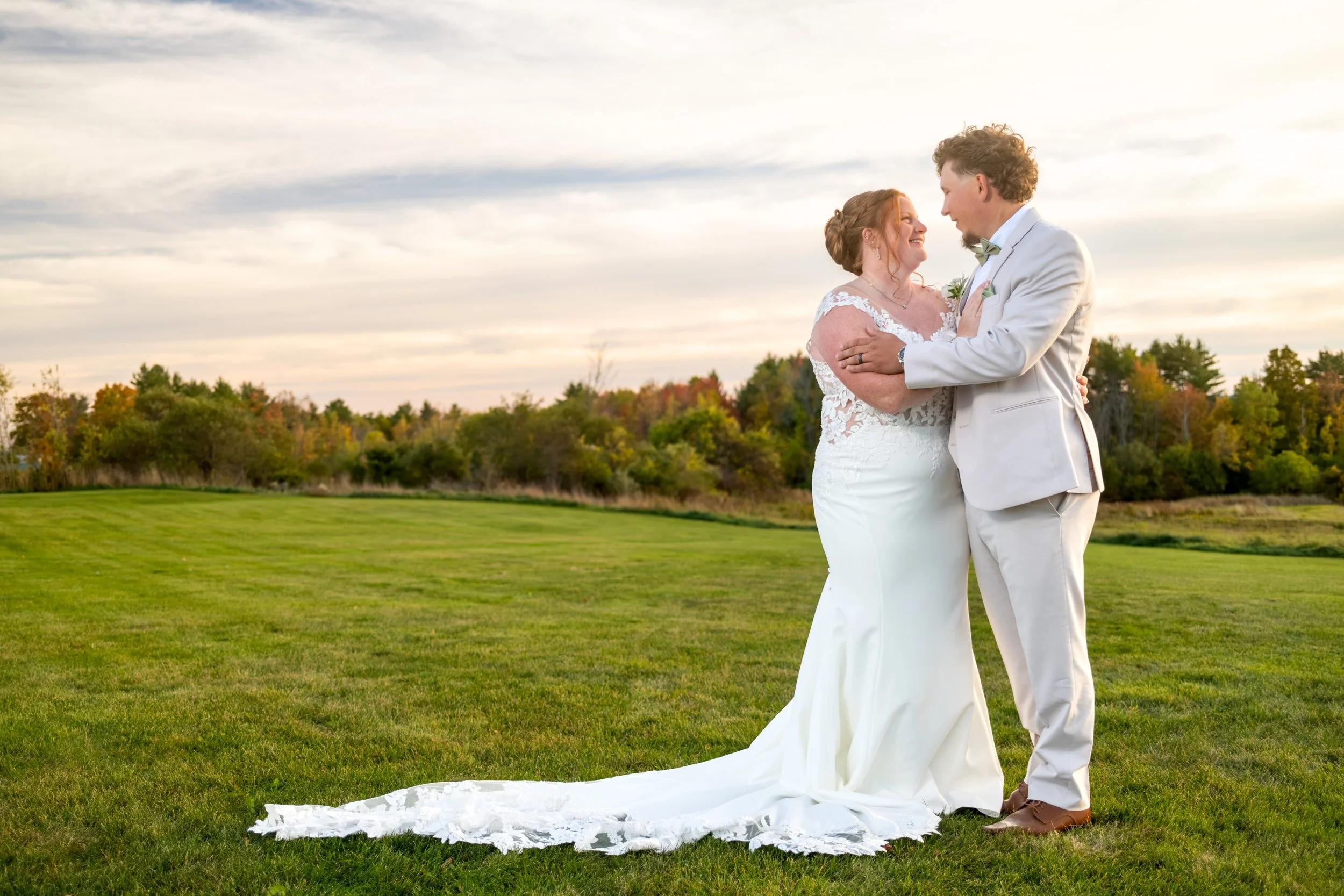 Erika and Andrew embracing in the field with her train spread across the grass at the Maine Wedding Barn and Event Center in Minot Maine fall wedding photography VMG Productions