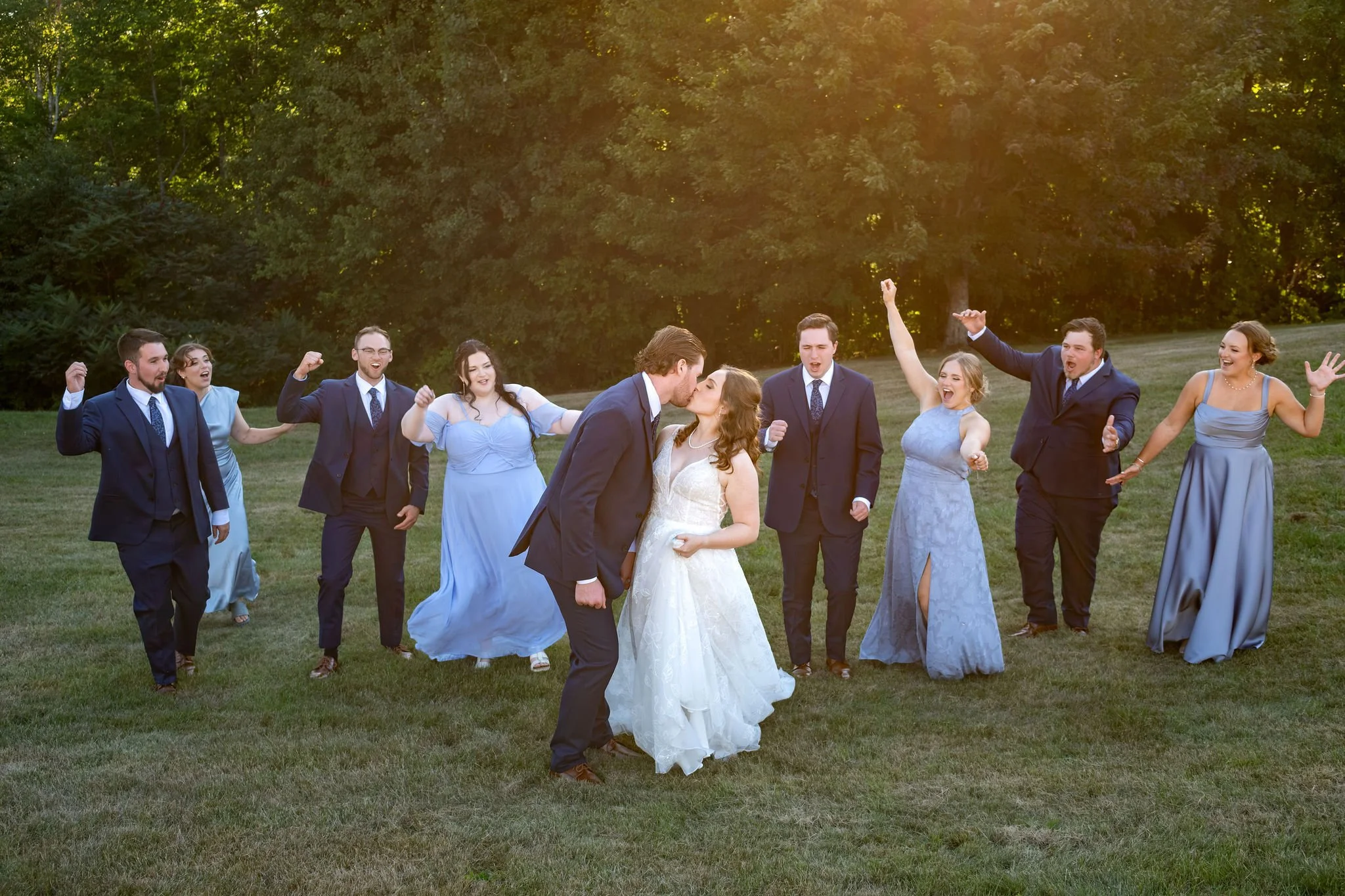Zachary and Hollie kissing in a field while their wedding party cheers on either side in golden light at The Lucerne Inn in Dedham Maine summer wedding photography VMG Productions