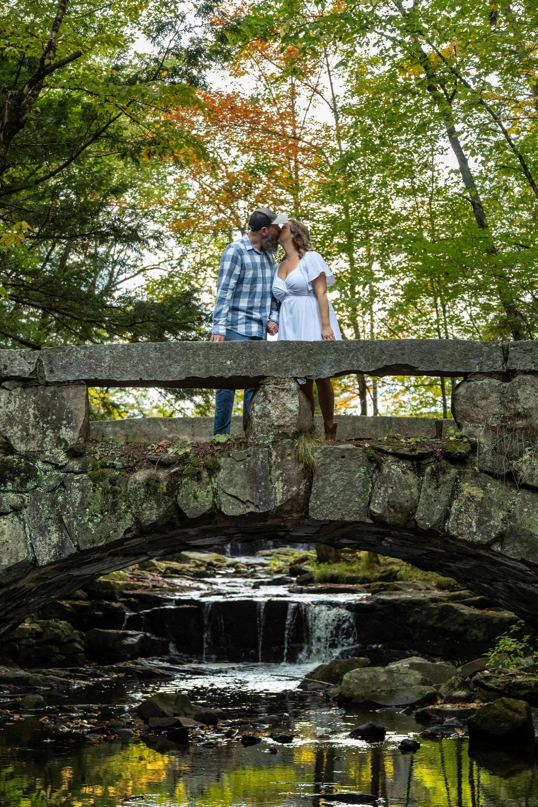 Alison and Dave sitting on top of the granite arch bridge with the waterfall and the pool below and orange leaves above at Vaughan Woods in Hallowell Maine fall engagement photography VMG Productions