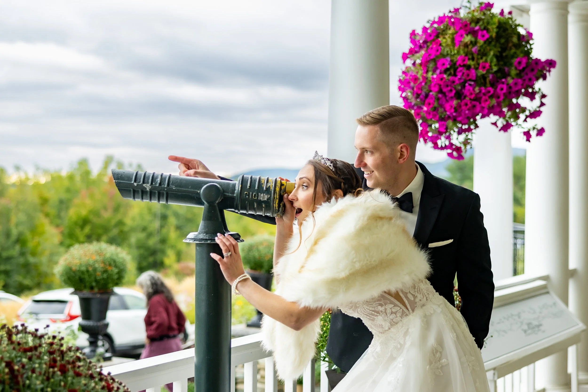 Bride and groom laughing together on grand hotel porch with mountain views and hanging fuchsia flower baskets at Mountain View Grand Resort Whitefield New Hampshire fall wedding 2022