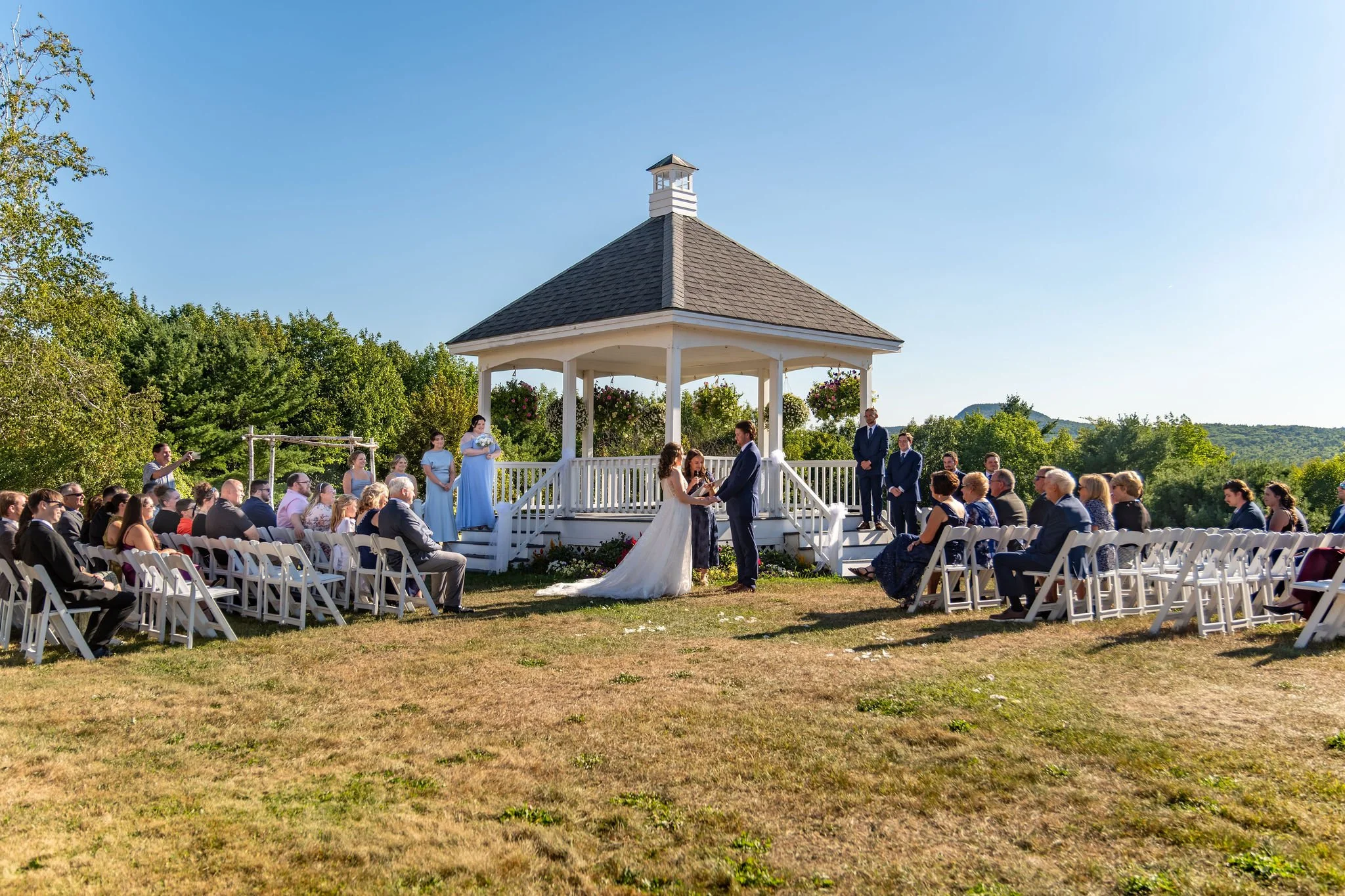 Wide view of Zachary and Hollie's ceremony at the hilltop gazebo with guests seated on the lawn under a blue sky at The Lucerne Inn in Dedham Maine summer wedding photography VMG Productions