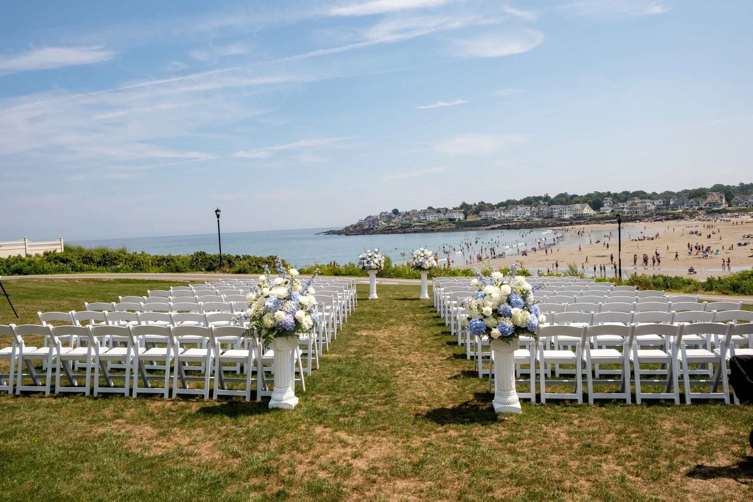 White ceremony chairs and blue hydrangea arrangements set on the oceanfront lawn above the crowded beach before the wedding at Union Bluff Hotel in York Maine wedding photography VMG Productions