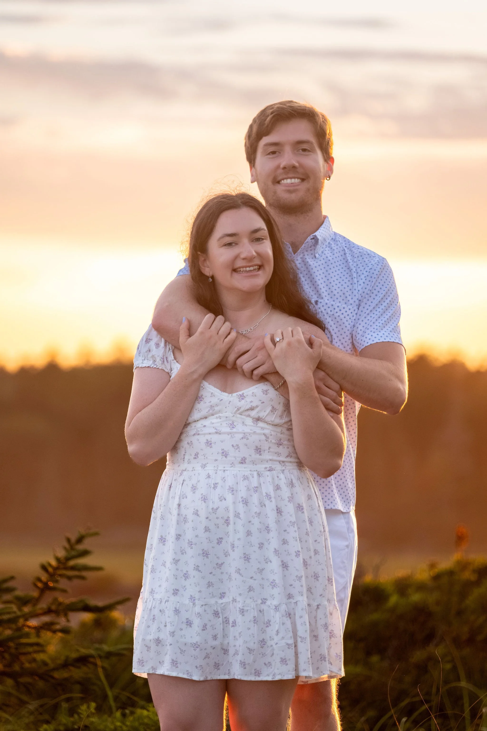Lucas wrapping his arms around Kirstie from behind with the sunset glow over the marsh at Reid State Park in Georgetown Maine engagement photography VMG Productions