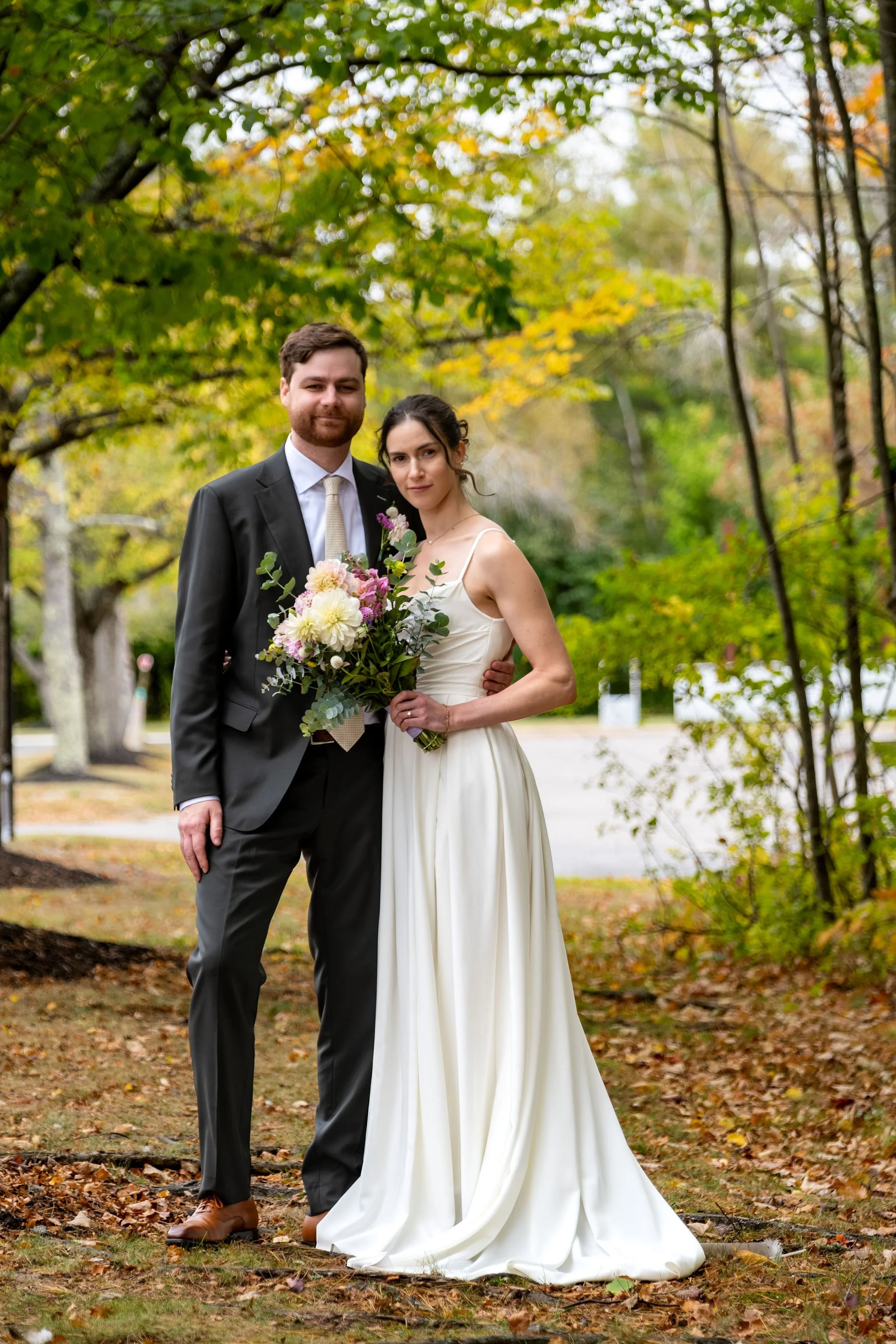 Victoria and Jack posing in fallen leaves beneath early fall foliage at The Cellars at Allagash Brewing Company in Portland Maine fall wedding photography VMG Productions