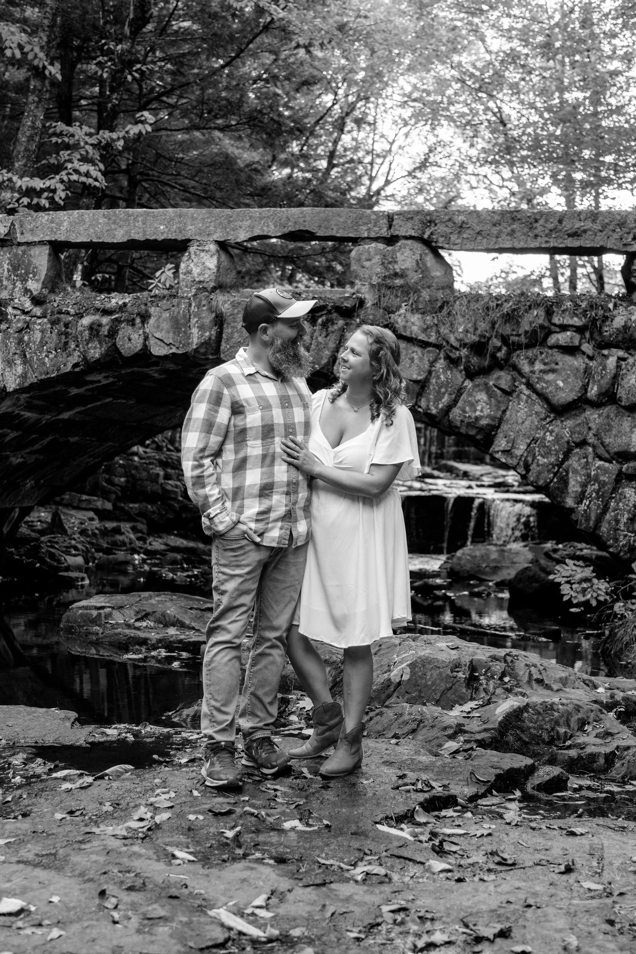 Black and white of Alison and Dave looking at each other below the arch bridge at Vaughan Woods in Hallowell Maine fall engagement photography VMG Productions