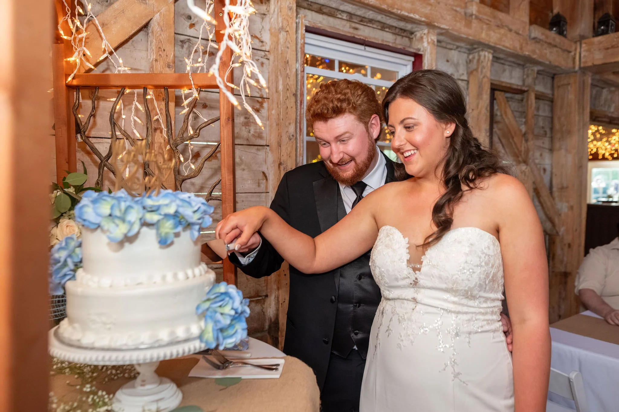 Alyssa and Colby cutting their wedding cake decorated with blue hydrangeas at the 1888 Barn Bethel Maine summer wedding photography VMG Productions