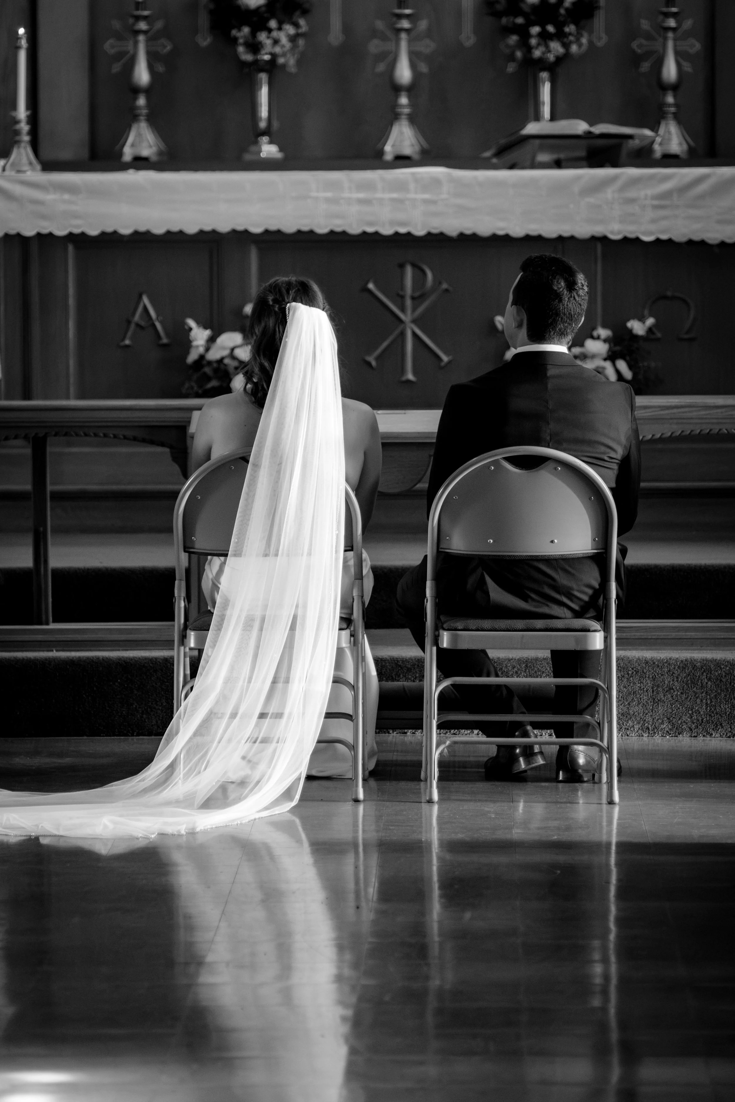 Caroline and Anthony Pirolli seated at the altar from behind with the cathedral veil pooling on the floor in black and white at Star of the Sea in York Maine wedding photography VMG Productions