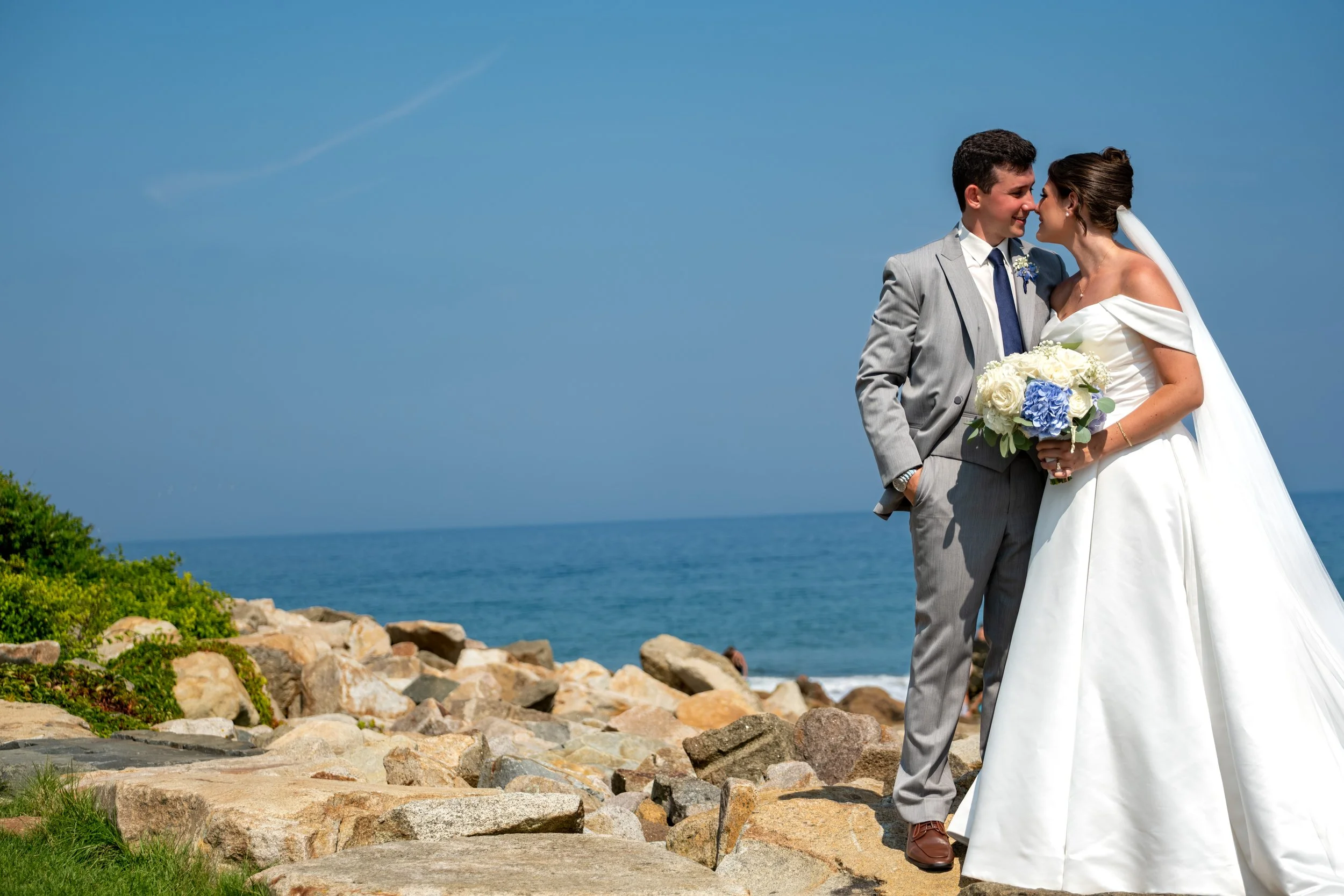 Caroline and Anthony Pirolli nose to nose on the rocks above the beach with the open Atlantic and summer sky behind them at Union Bluff Hotel in York Maine wedding photography VMG Productions