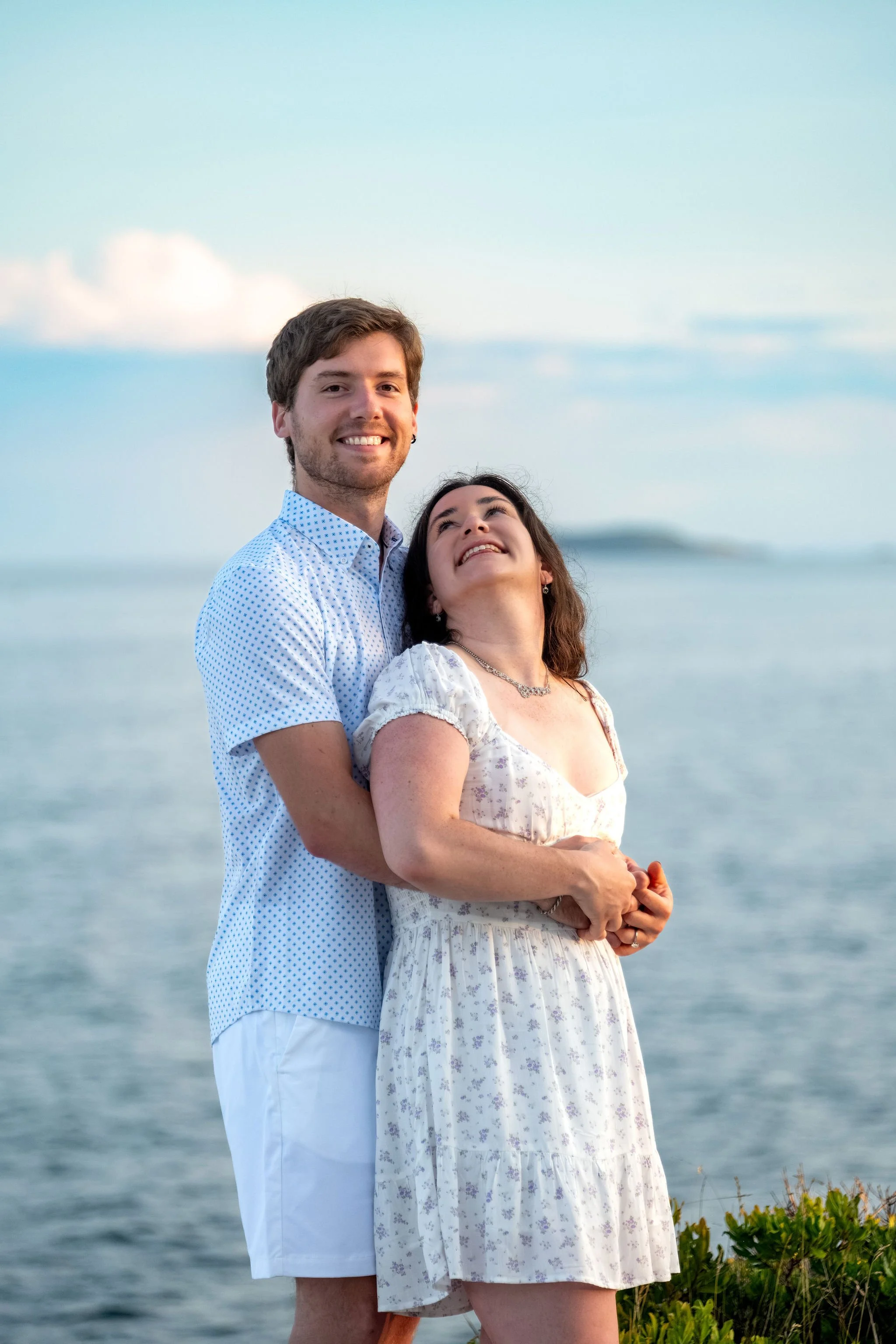 Lucas holding Kirstie from behind with the ocean and an island on the horizon at Reid State Park in Georgetown Maine engagement photography VMG Productions