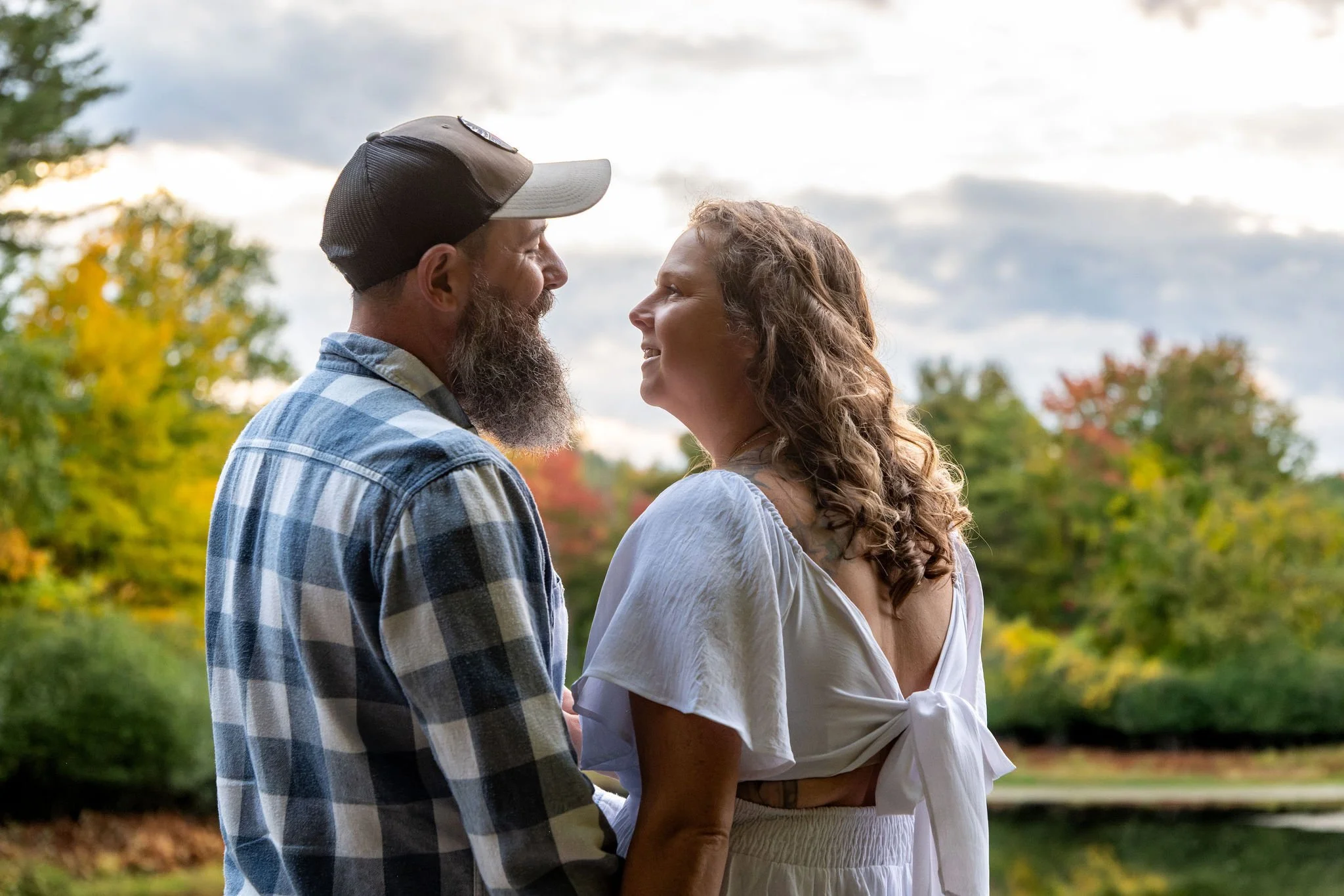 Alison and Dave laughing at each other at the edge of the pond with the fall trees behind them at Vaughan Woods in Hallowell Maine fall engagement photography VMG Productions