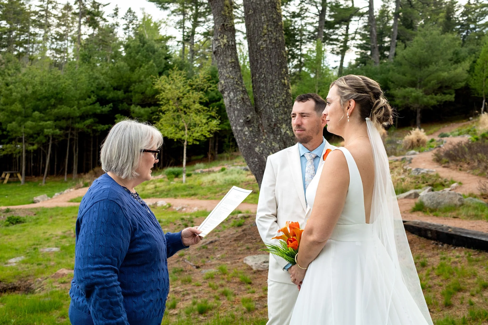 The officiant reading vows to Alli and Adam Baudoin under the pines at their private residence ceremony in Mount Desert Maine spring wedding photography VMG Productions