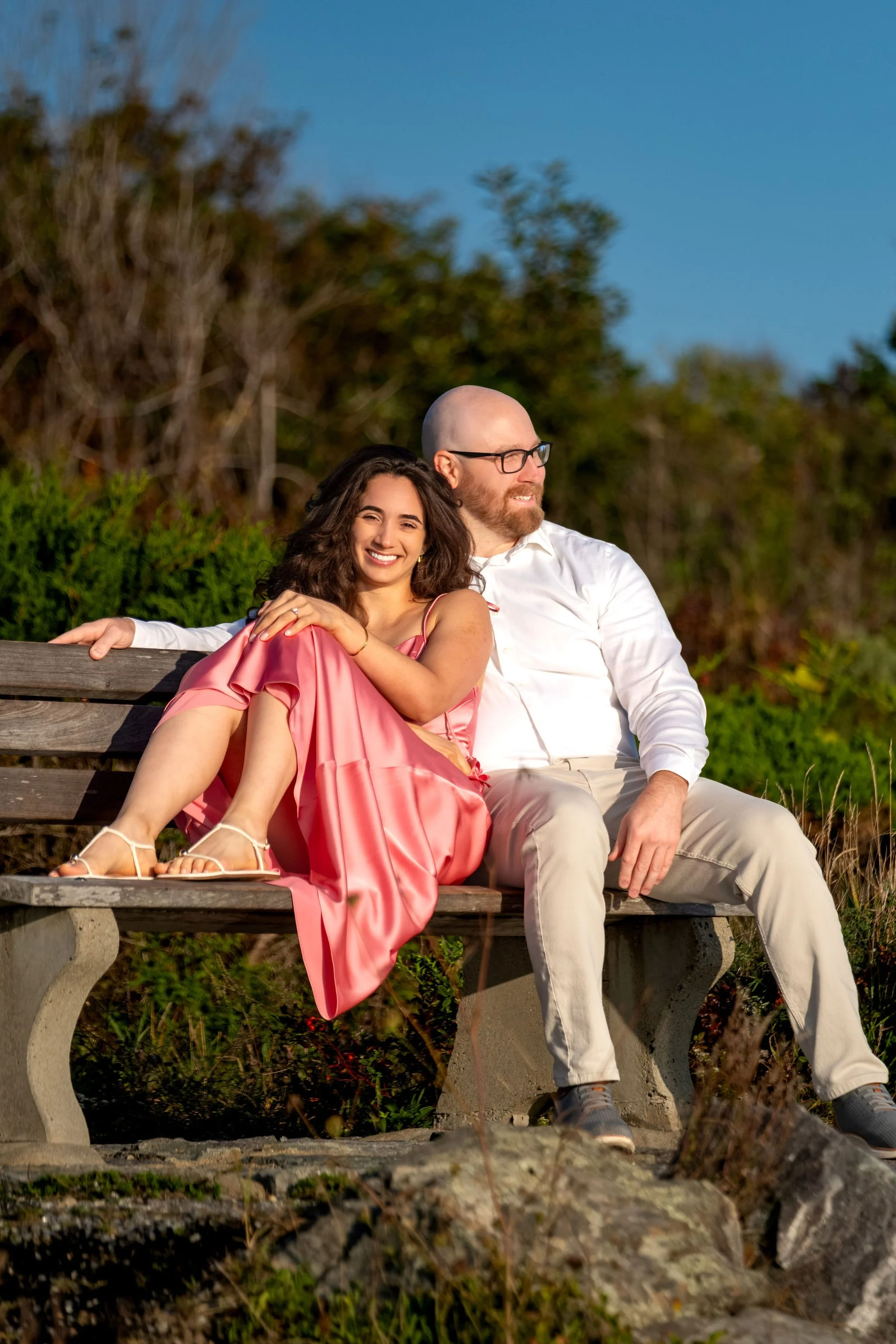 Helena leaning back against Zack on a weathered bench with the coastal shrubs behind them on Marginal Way in Ogunquit Maine sunrise proposal and engagement photography VMG Productions