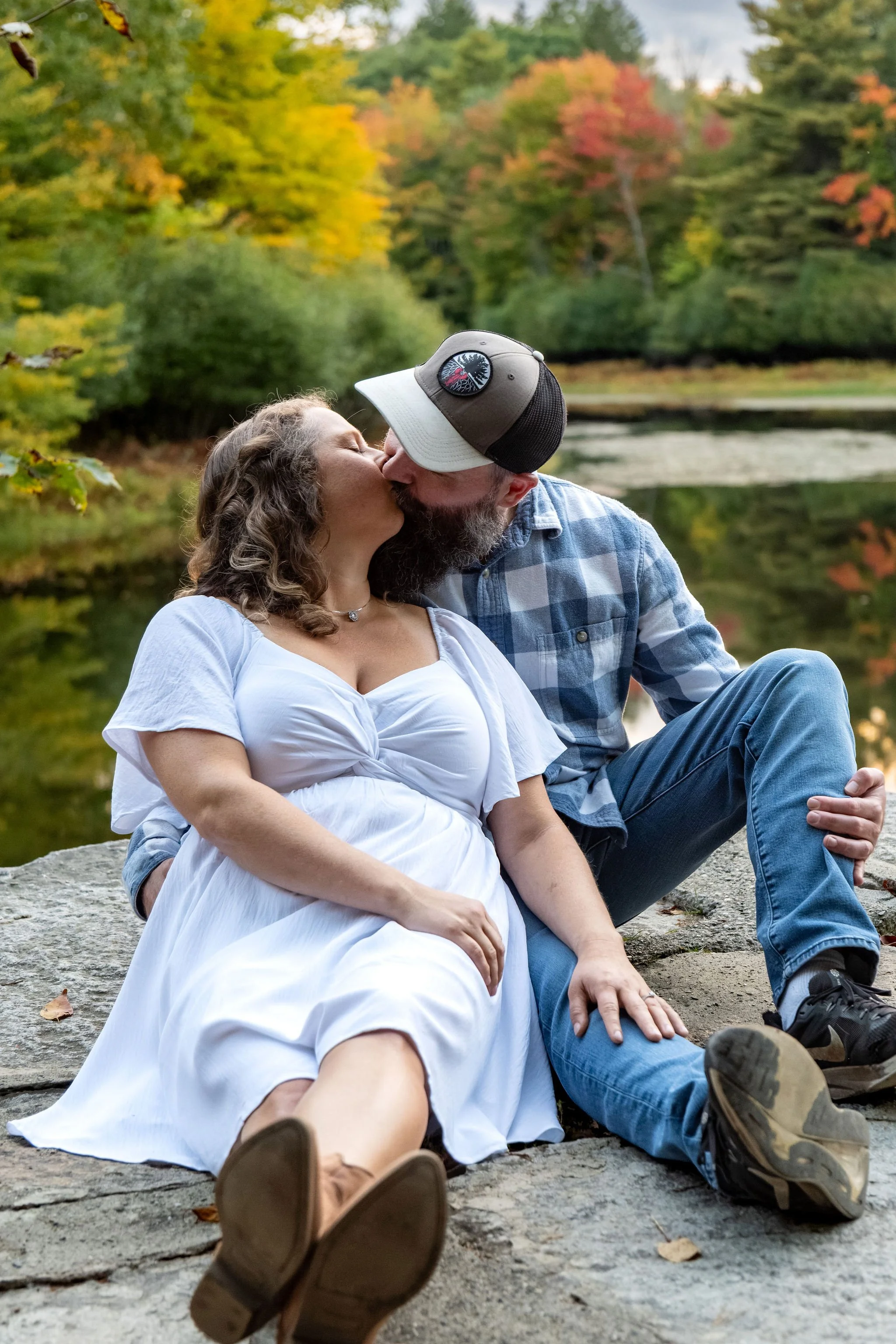 Alison and Dave kissing while sitting on the rocks at the pond with the fall color behind them at Vaughan Woods in Hallowell Maine fall engagement photography VMG Productions