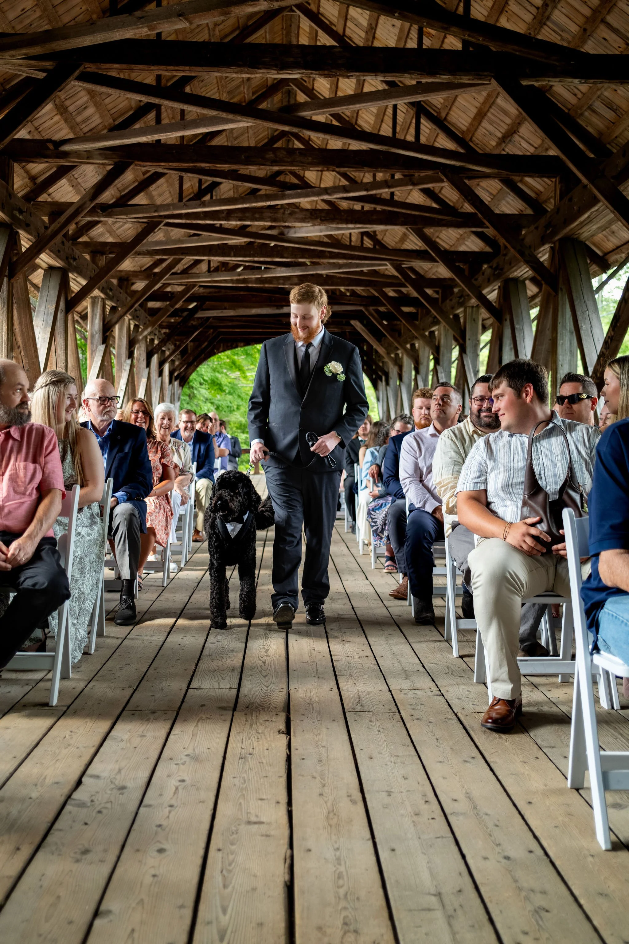 Alyssa and Colby the groom walking Ryder the dog down the aisle as guests smile at Sunday River Covered Bridge Newry Maine summer wedding photography VMG Productions
