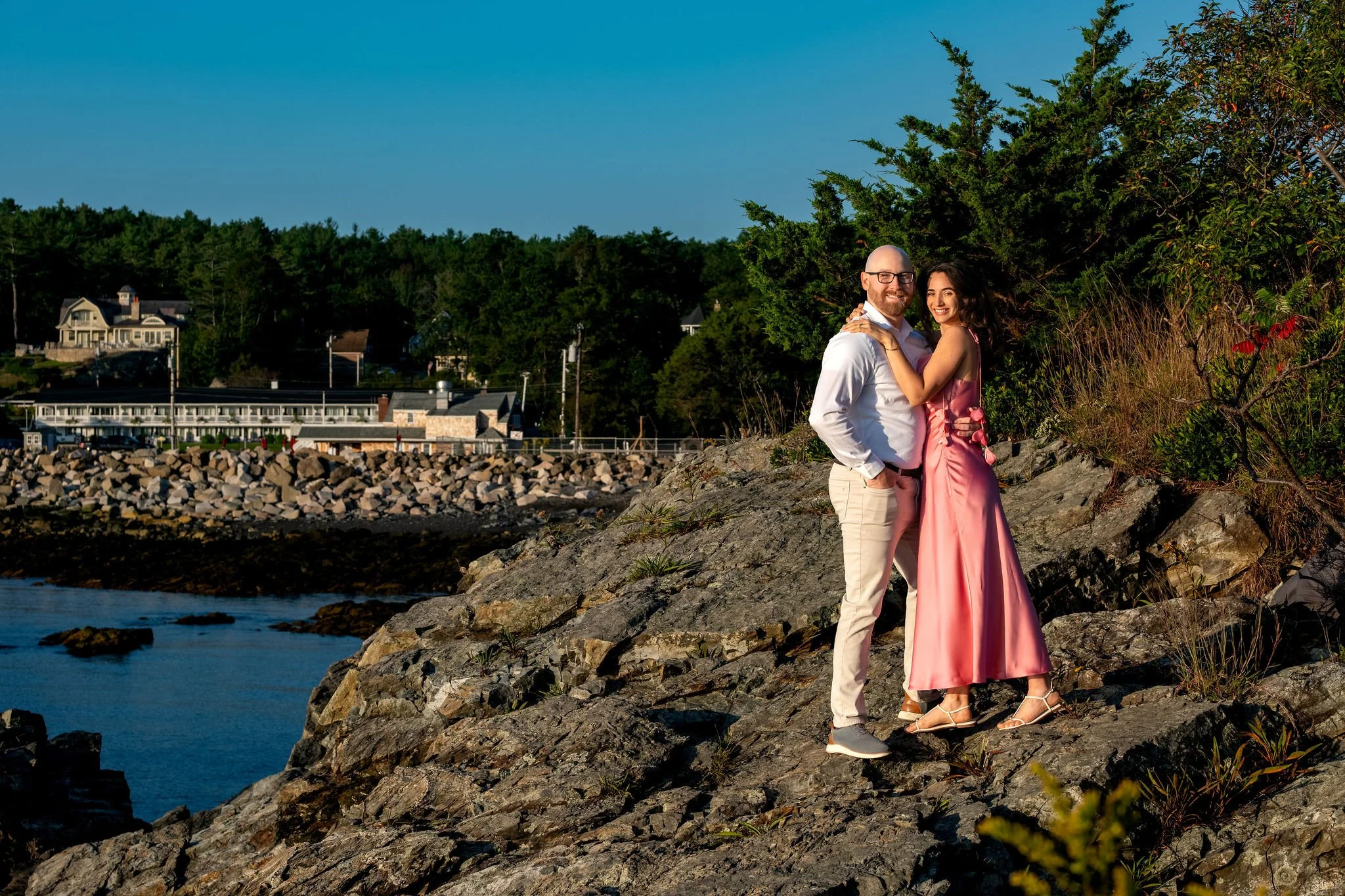 Zack and Helena standing together on a rocky ledge above the water with the harbor in the distance on Marginal Way in Ogunquit Maine sunrise proposal and engagement photography VMG Productions