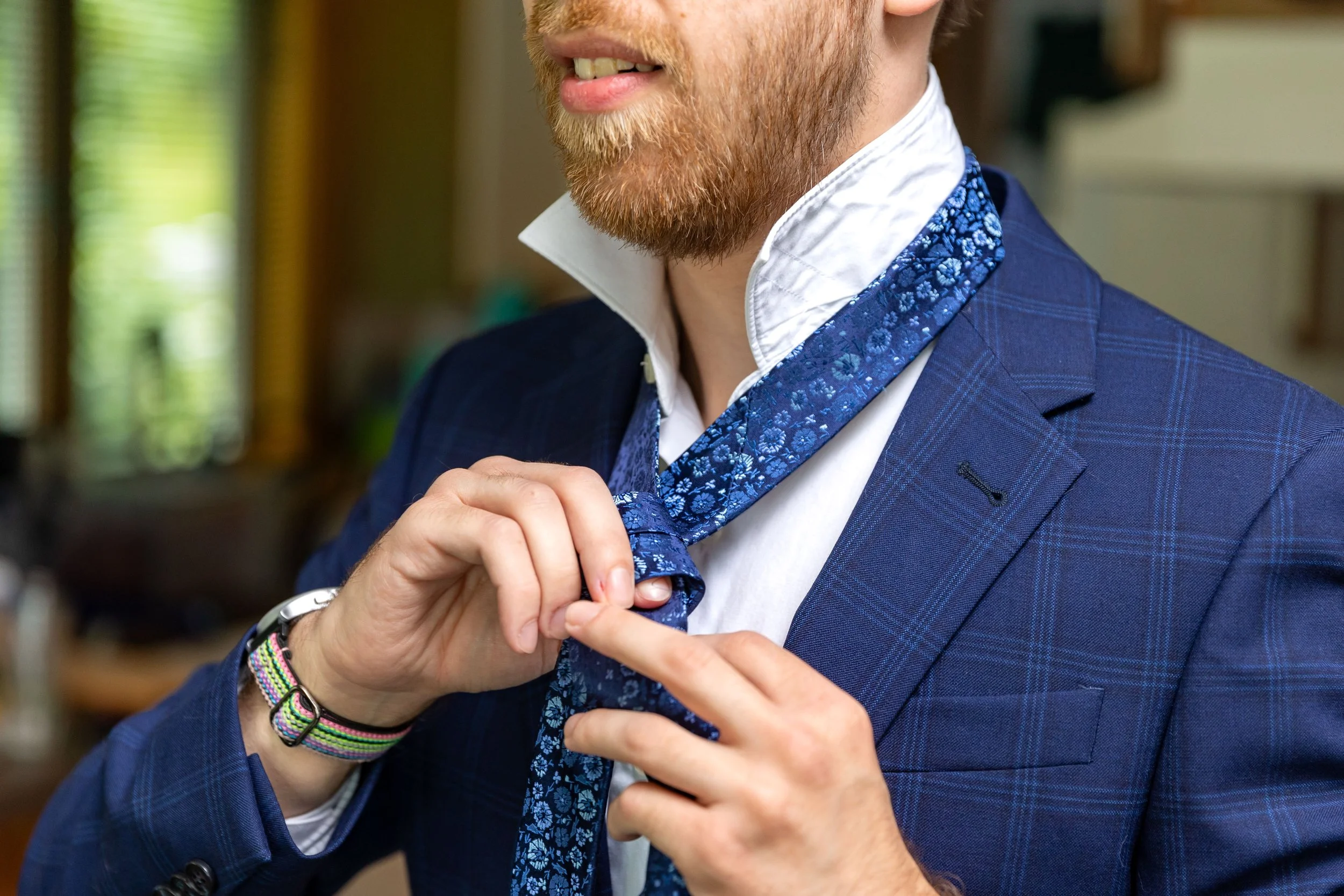 Groom with red beard tying blue floral patterned necktie on navy blue plaid suit jacket during wedding morning getting ready at Owls Head Maine coastal wedding 2024