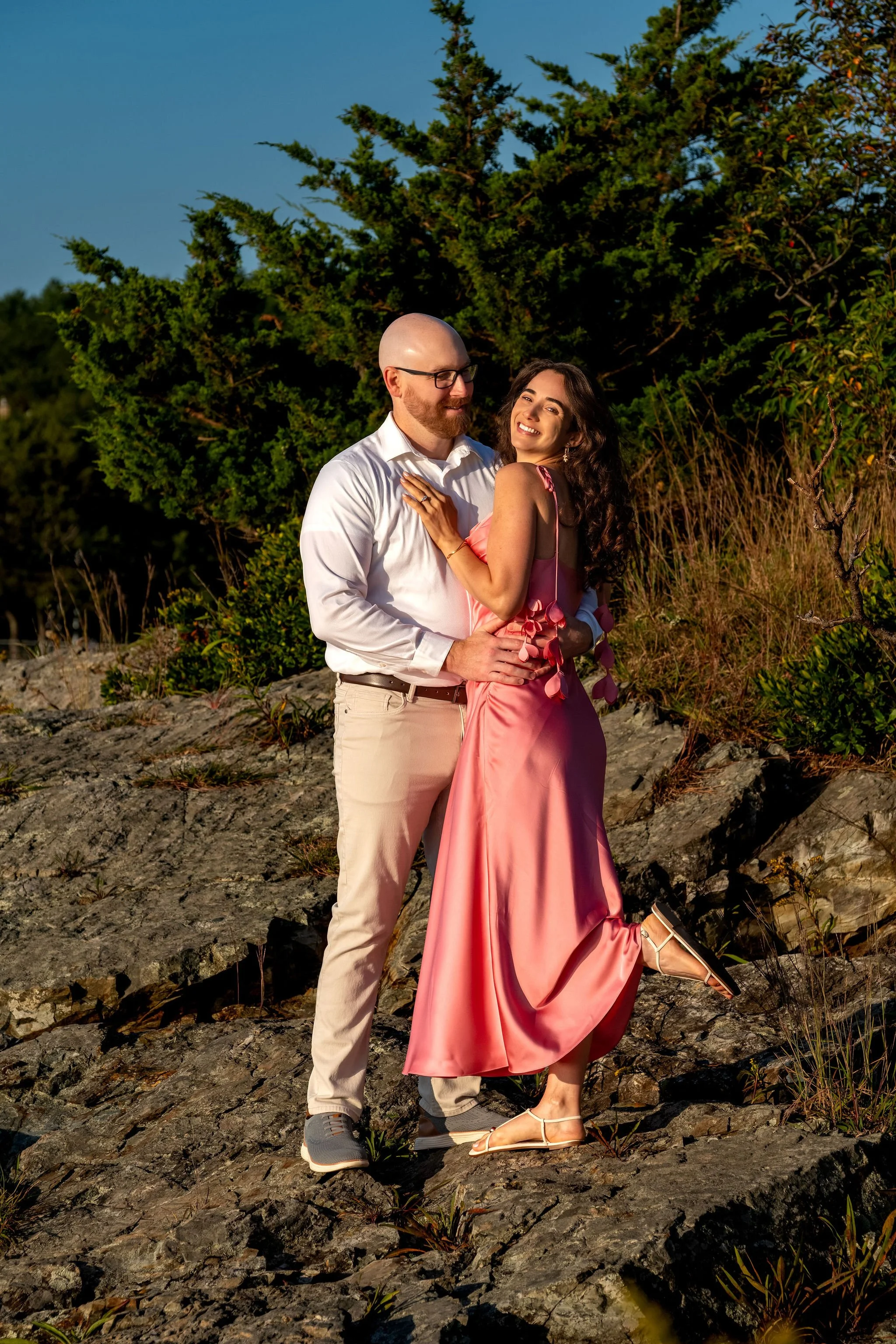Helena laughing over Zack's shoulder as they hug on the rocks with her foot kicked up on Marginal Way in Ogunquit Maine sunrise proposal and engagement photography VMG Productions