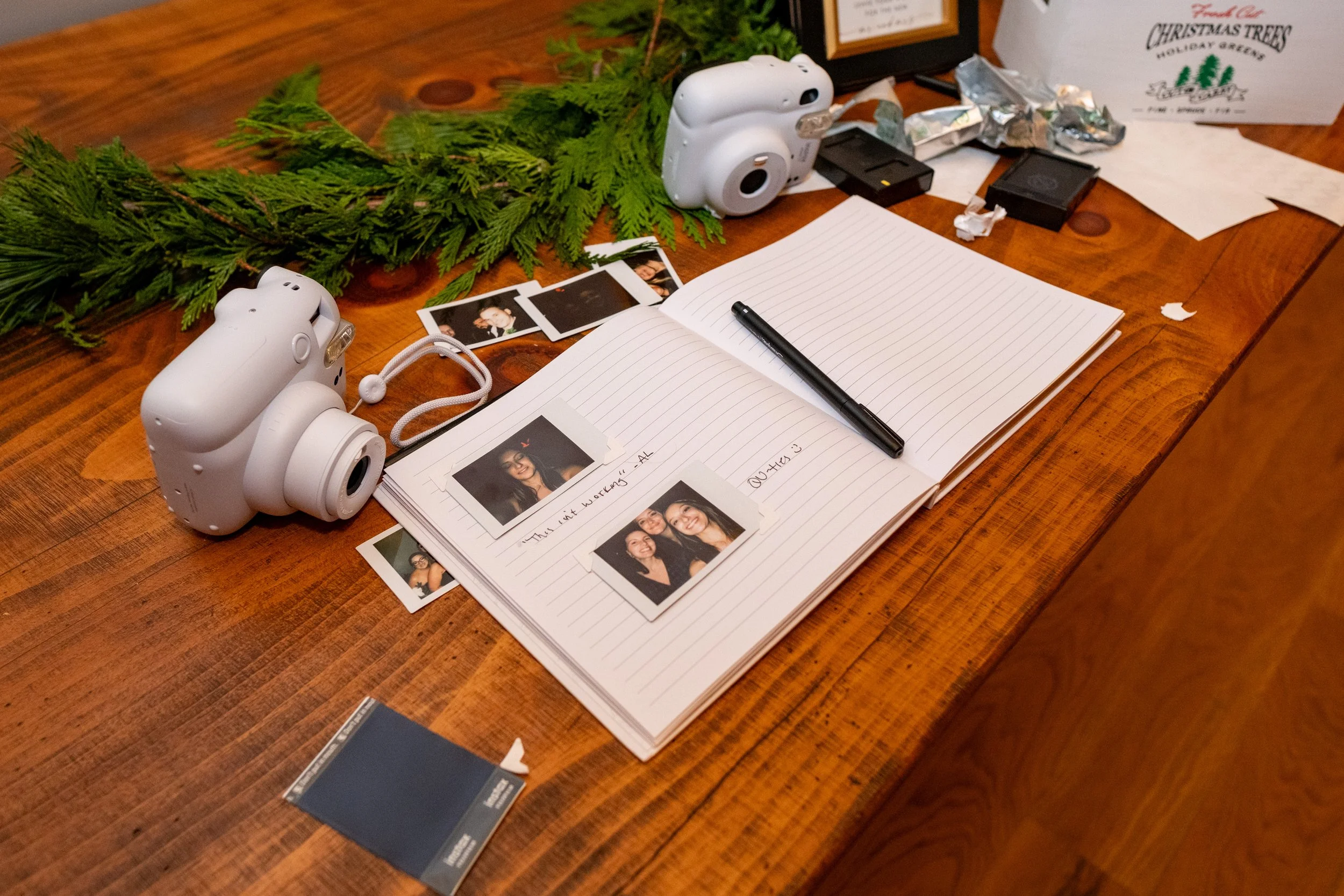 Instax camera guest book station at Old Saco Inn Fryeburg Maine winter wedding with open photo album showing polaroid prints and handwritten guest notes beside white Fujifilm cameras and cedar garland table decor