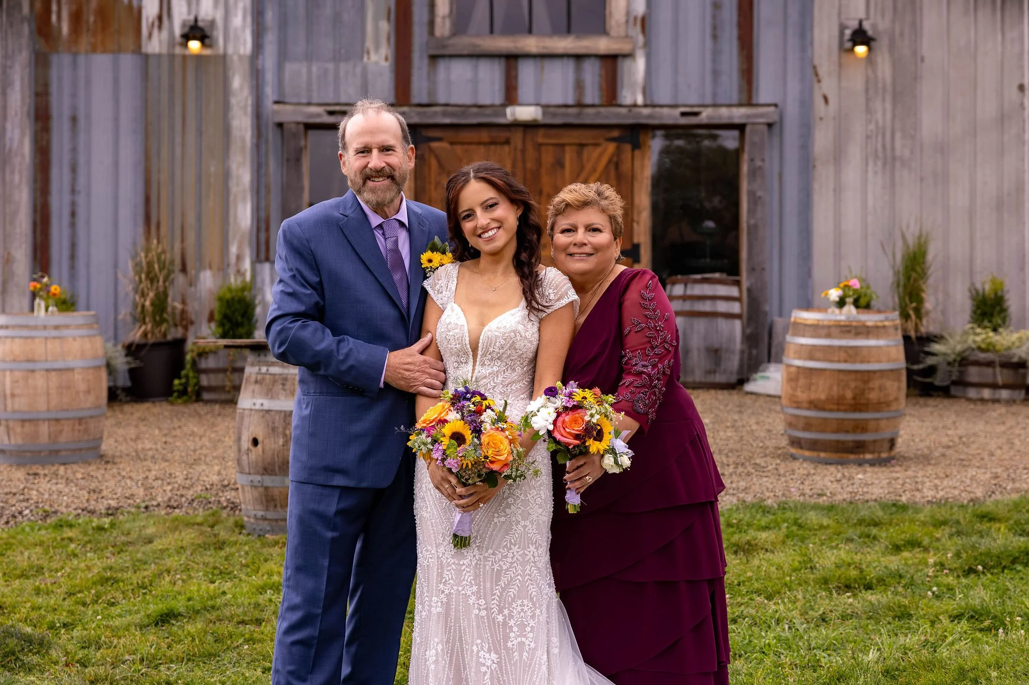 Bride Britney in beaded lace wedding gown with father in blue suit and mother in burgundy dress intimate three-person family formal portrait in front of weathered barn with wine barrels at Mountain Star Estate Rangeley Maine summer 2025 wedding
