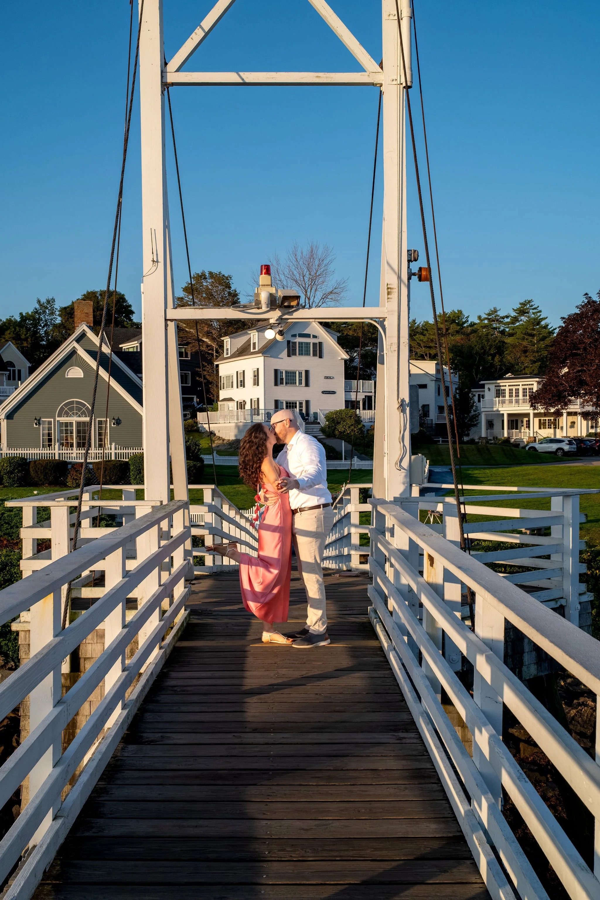 Zack and Helena kissing in the middle of the footbridge framed by the white railings on Marginal Way in Ogunquit Maine sunrise proposal and engagement photography VMG Productions