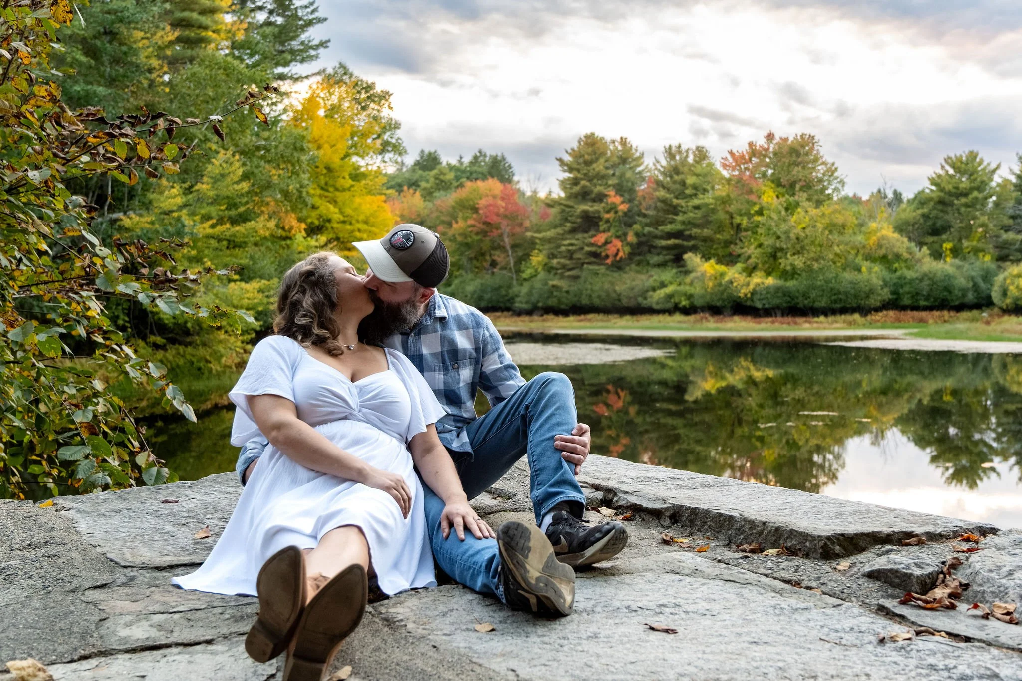 Alison and Dave sitting on the granite ledge and kissing with the pond and the clouds stretching out behind them at Vaughan Woods in Hallowell Maine fall engagement photography VMG Productions