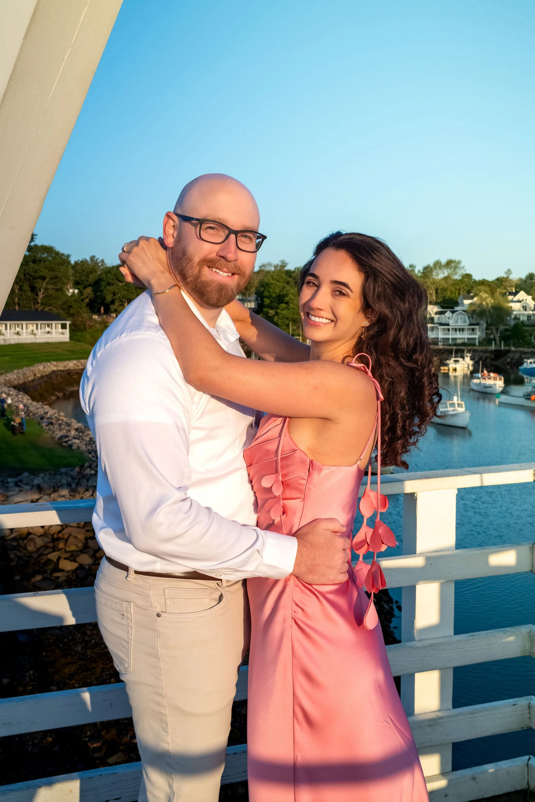 Helena beaming at the camera in Zack's arms with the white bridge beam and harbor behind them on Marginal Way in Ogunquit Maine sunrise proposal and engagement photography VMG Productions