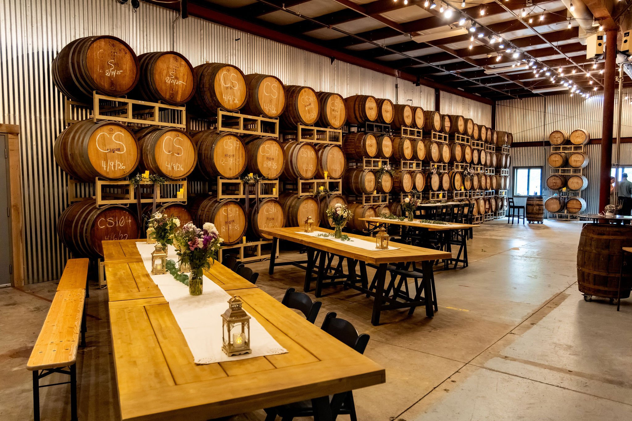 Wide view of the barrel room set with long wooden tables, lanterns, and string lights before guests arrived at The Cellars at Allagash Brewing Company in Portland Maine fall wedding photography VMG Productions
